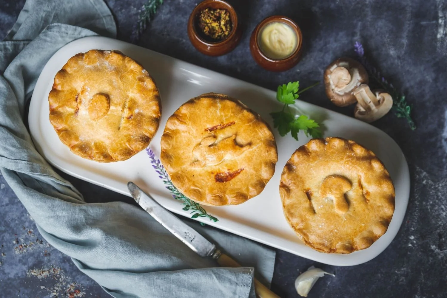 Three traditional baked pies on a white platter, accompanied by small bowls of condiments and garnished with herbs, set on a dark textured surface with a gray cloth and a small knife.