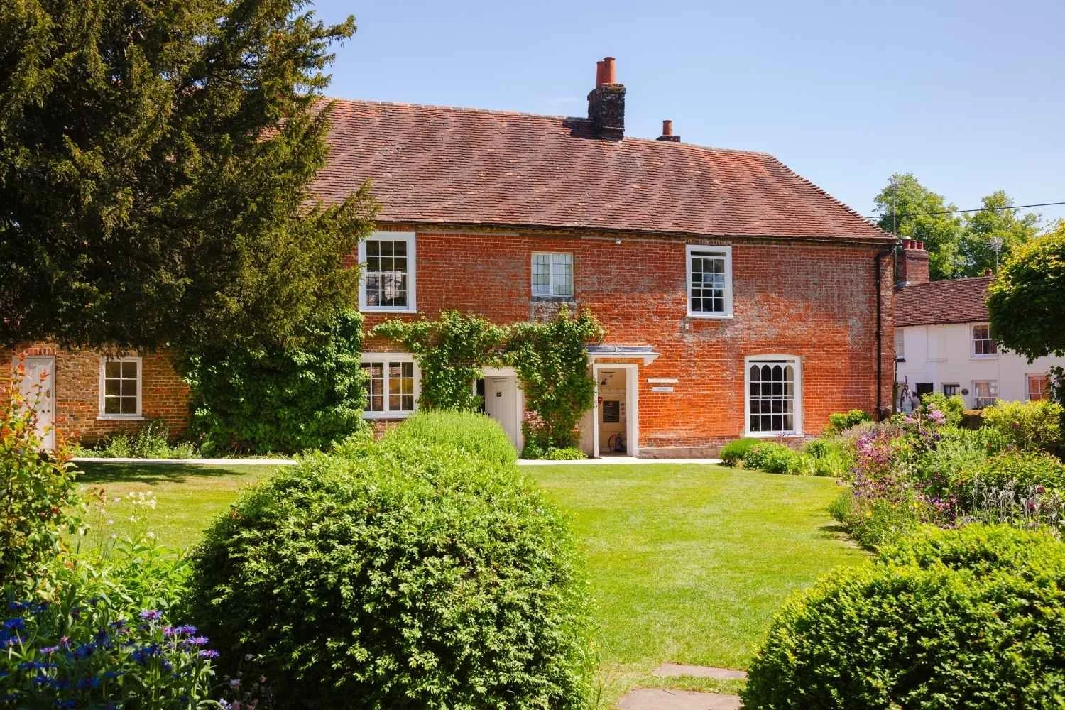 A red brick house with a brown tiled roof, surrounded by a lush green garden with bushes, trees, and flowering plants, under a clear blue sky.
