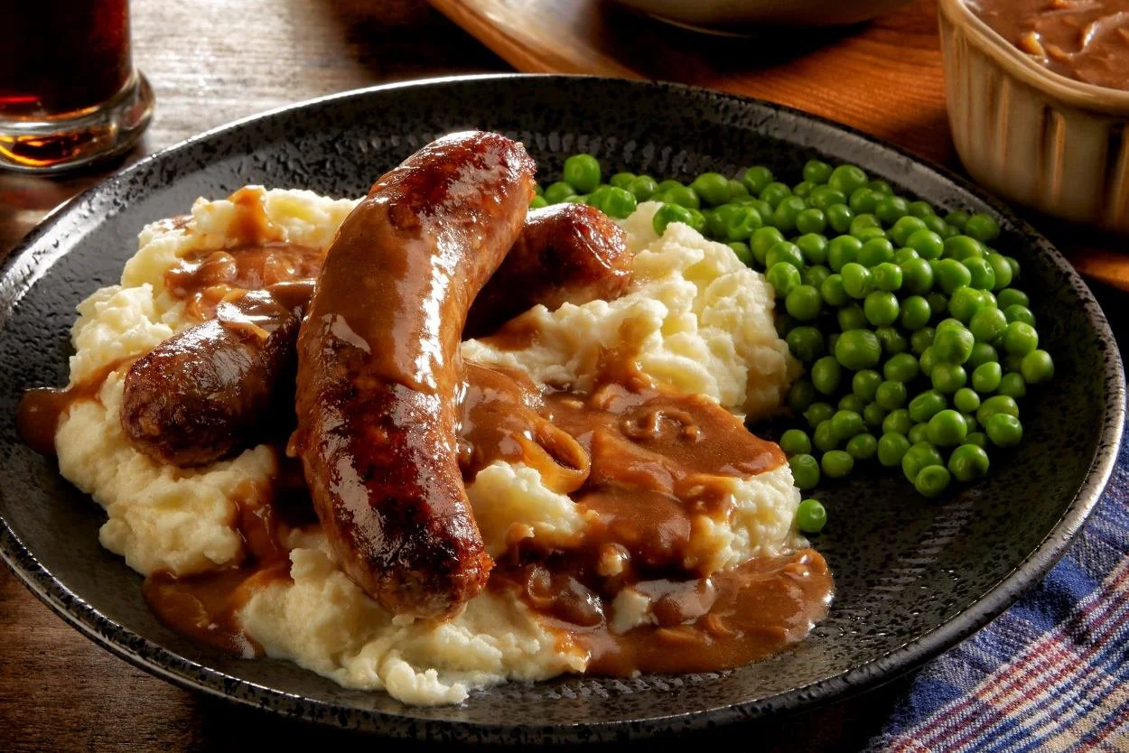 Plate of mashed potatoes with sausage links, brown gravy, green peas, and meatloaf in a bowl.