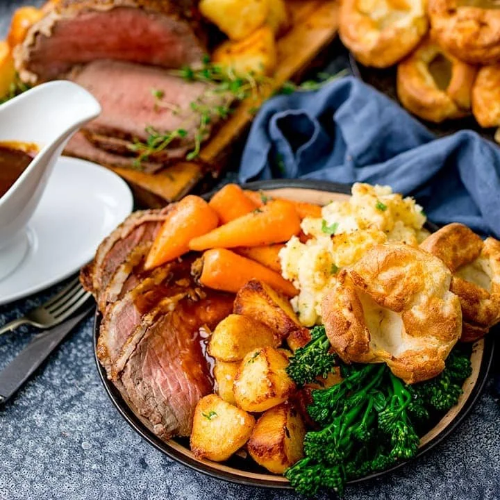 Plate of roasted prime rib with mashed potatoes, roasted carrots, broccoli, Yorkshire pudding, and gravy, surrounded by additional sliced prime rib and shrimp, with a side of gravy.