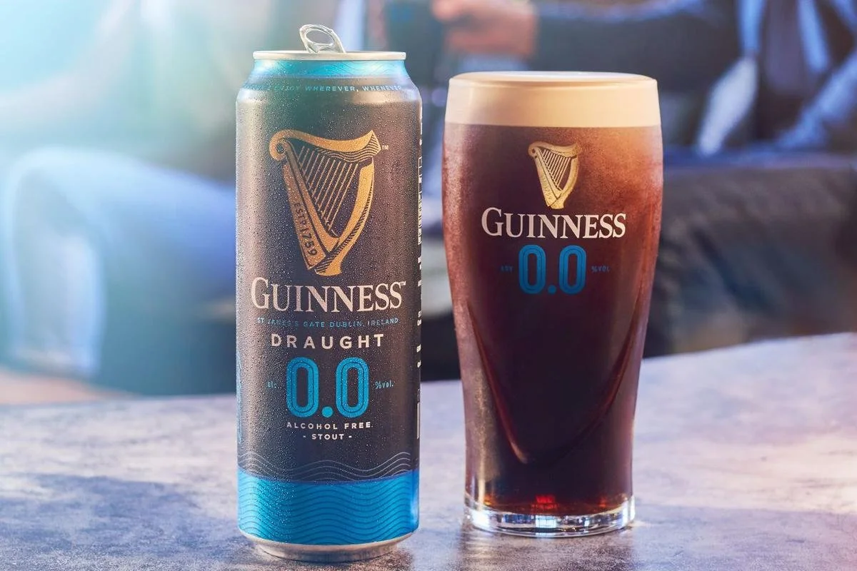 A can of Guinness Draught alcohol-free stout and a pint glass of the same with a dark stout, both with the Guinness logo and '0.0' on them, placed on a wooden surface.