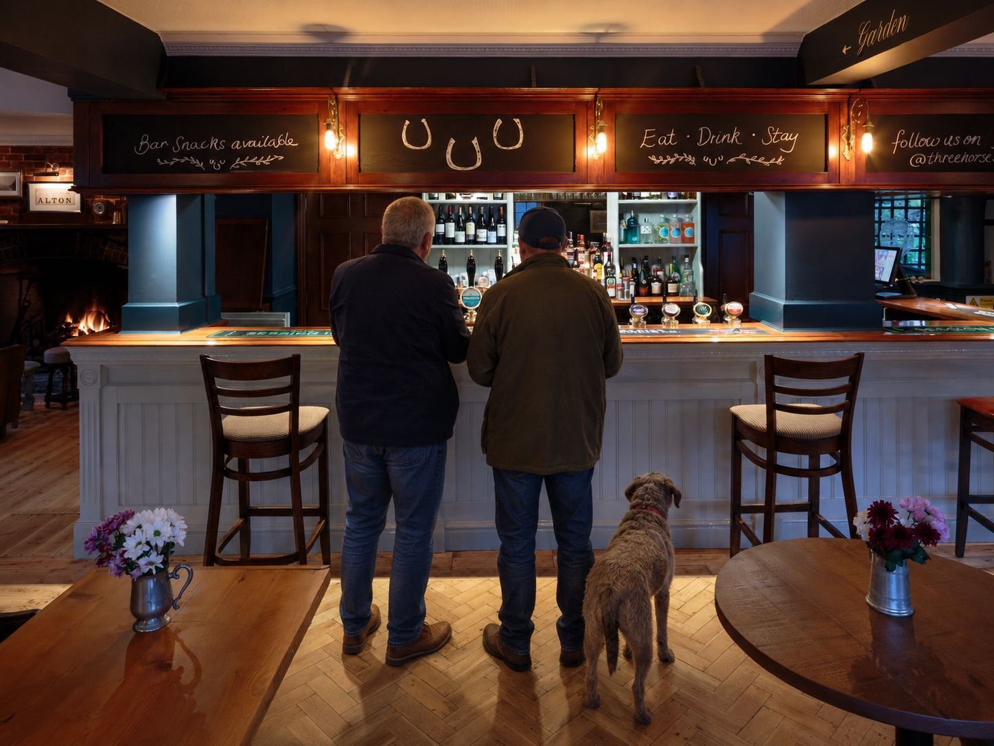 Two men and a dog standing at a bar counter, facing away from the camera, inside a cozy pub with bar shelves filled with bottles behind them. There are decorated tables with flower vases in the foreground and chalkboard signs with handwritten menus overhead.