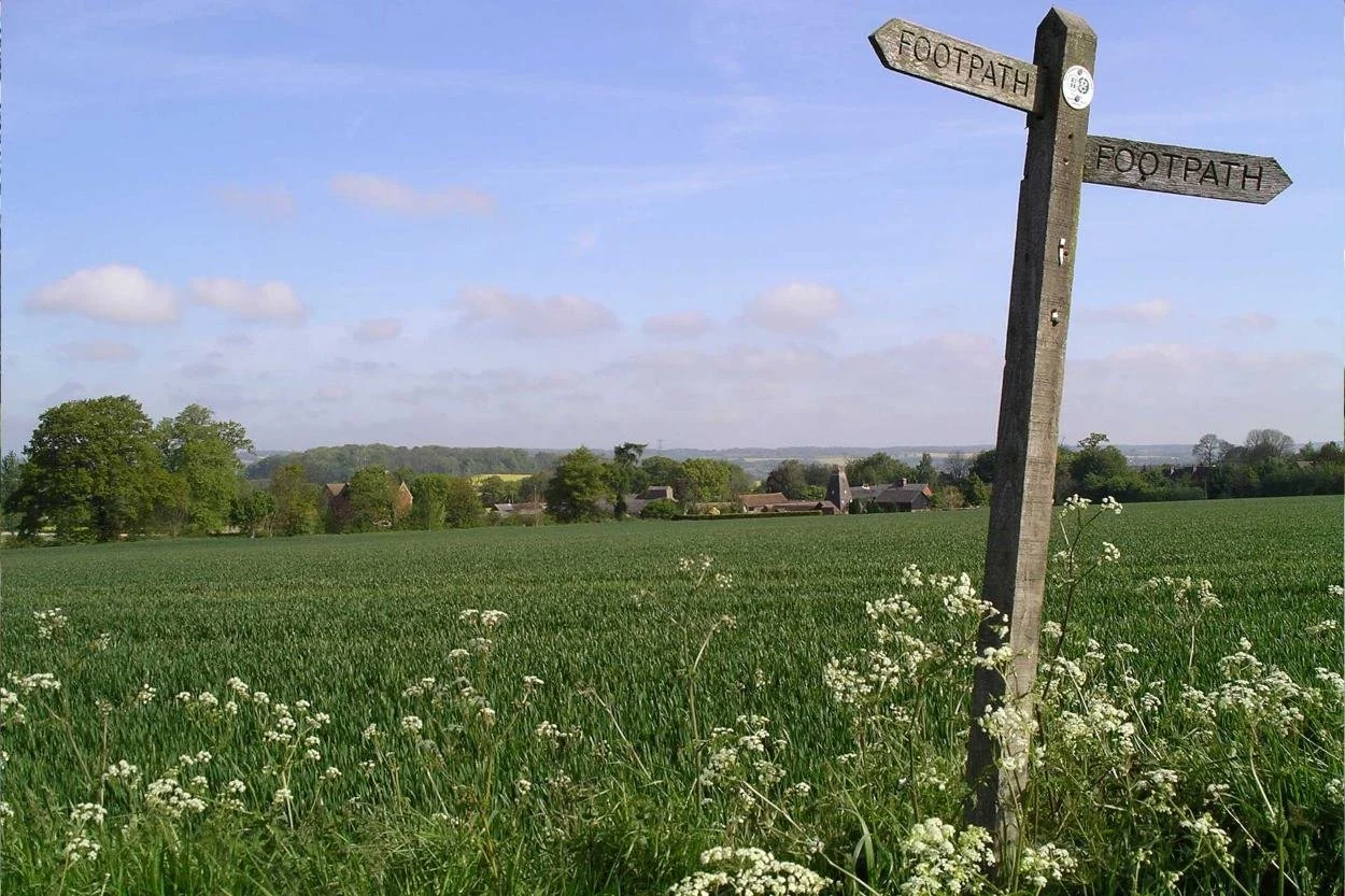 A countryside surrounded by green fields and trees under a partly cloudy sky, with a wooden signpost pointing in two directions labeled 'Footpath'.