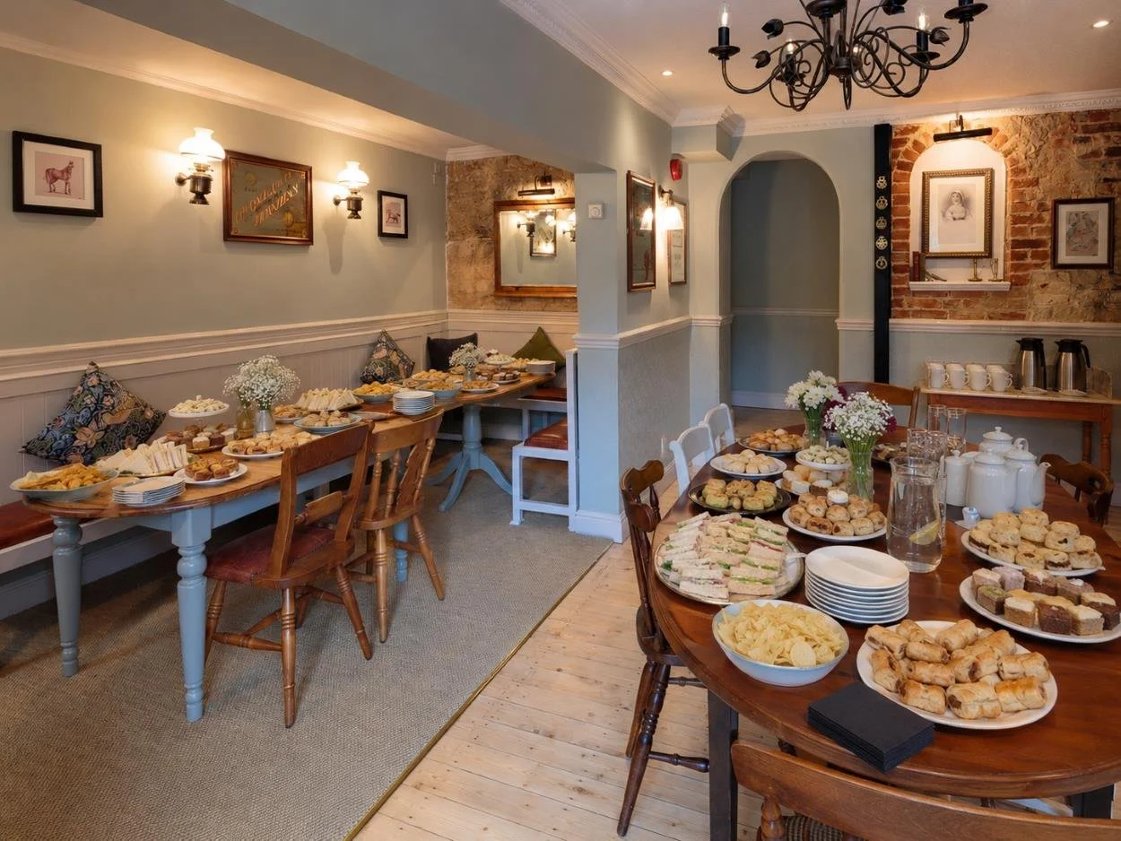 A room set up for a gathering with tables of assorted finger foods, snacks, and desserts, including sandwiches, cookies, chips, and pastries, with flowers, cups, and jugs on the tables, in a cozy, decorated space with framed art and warm lighting.