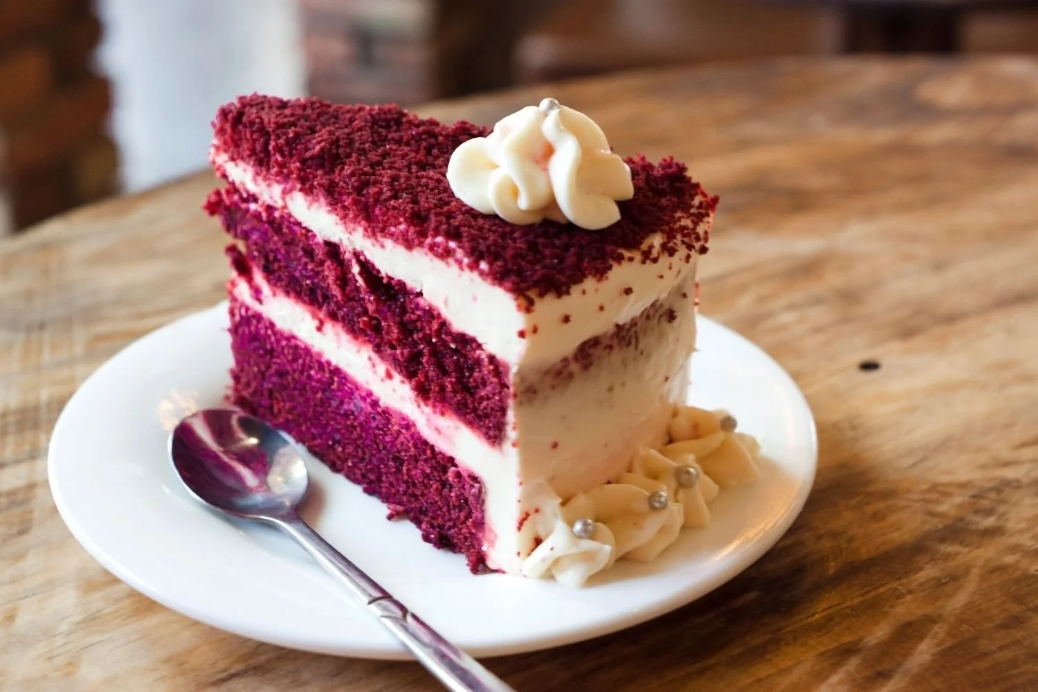 A slice of red velvet cake with cream cheese frosting, topped with white frosting dollop and red velvet crumbs, served on a white plate with a spoon.