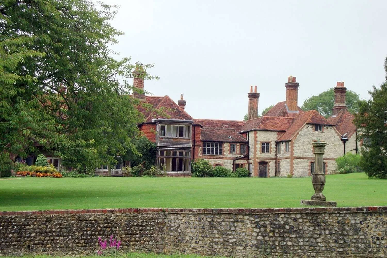 Historic brick and stone mansion with multiple chimneys, surrounded by a lush green lawn and trees. A stone wall runs across the foreground with decorative stone urns on the lawn.