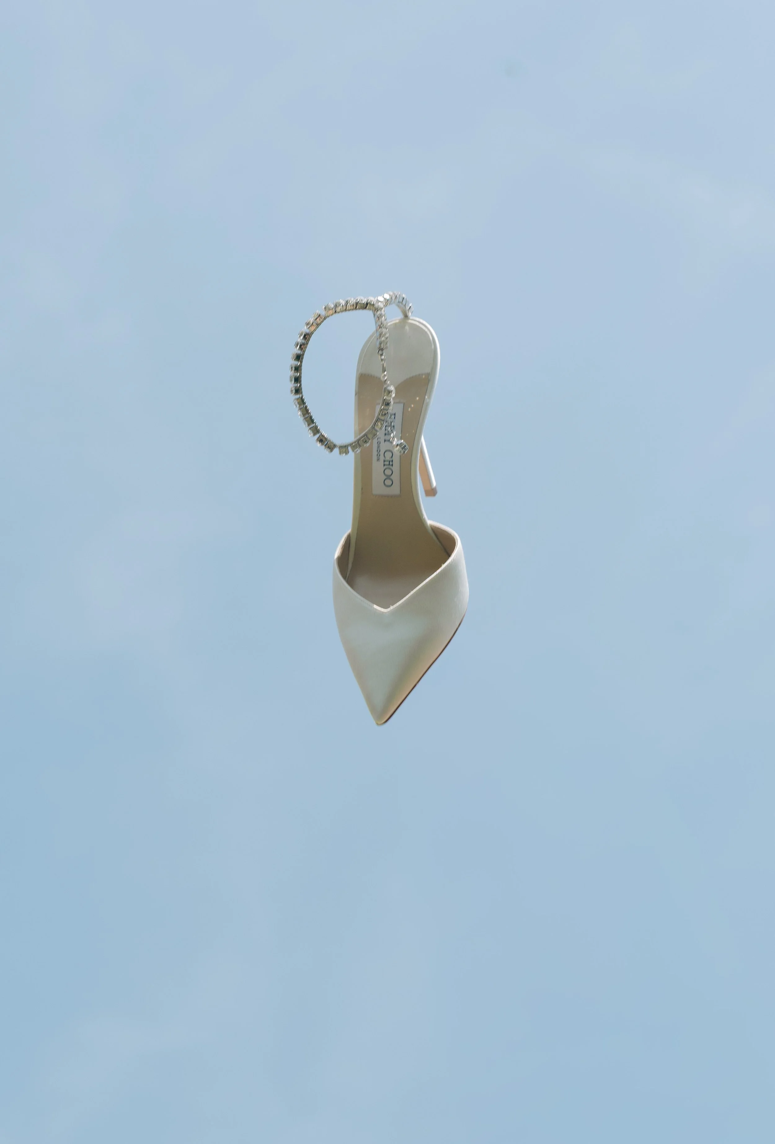 Weiße High-Heel-Schuhe, mit einer silbernen Schmuckkette und einem Anhänger, hängen an einem Draht in den Himmel gegen ein hellblaues Himmel