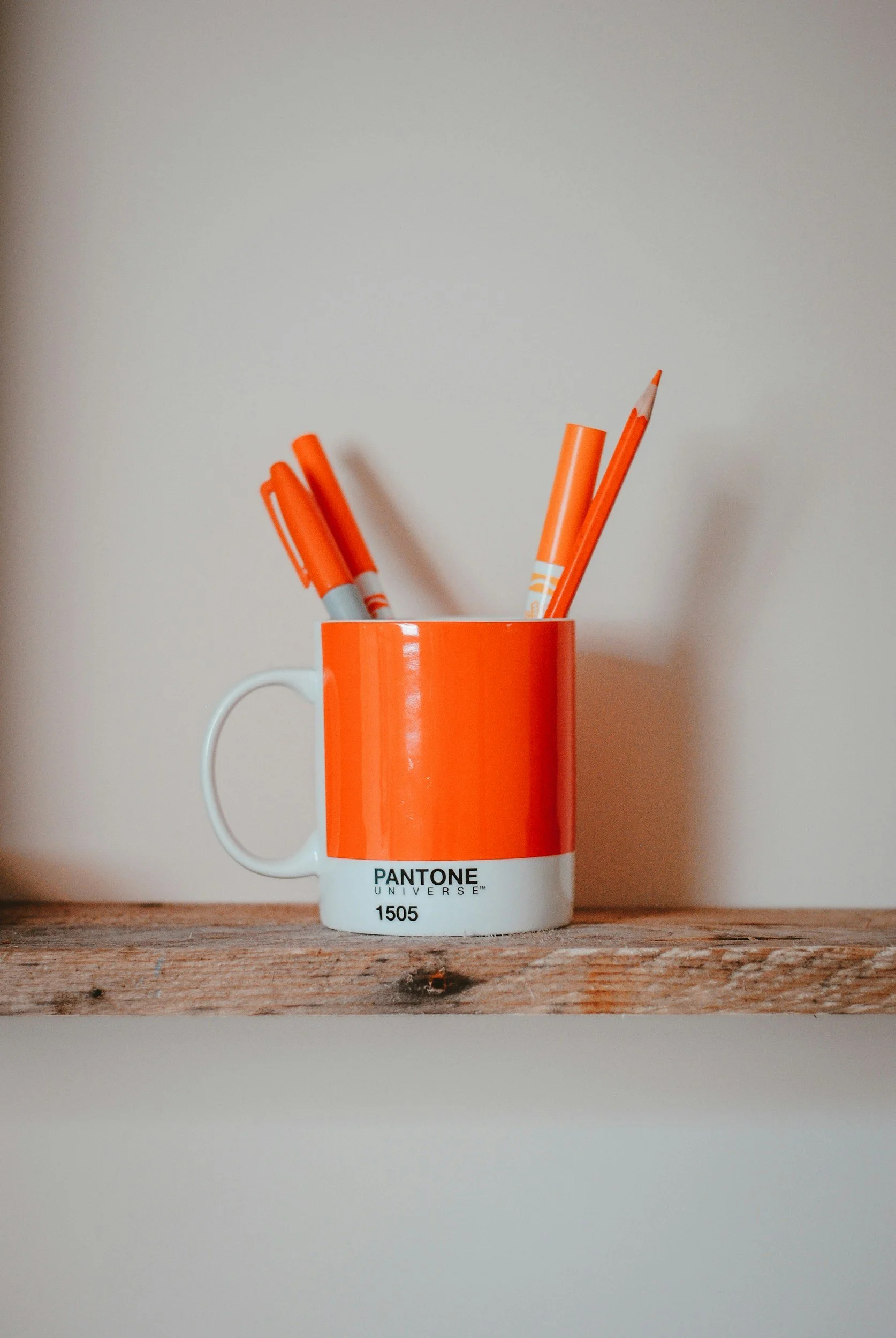 Orange and white mug labeled Pantone Universe, with orange markers inside, sitting on a wooden surface against a plain wall background.  Depicts branding