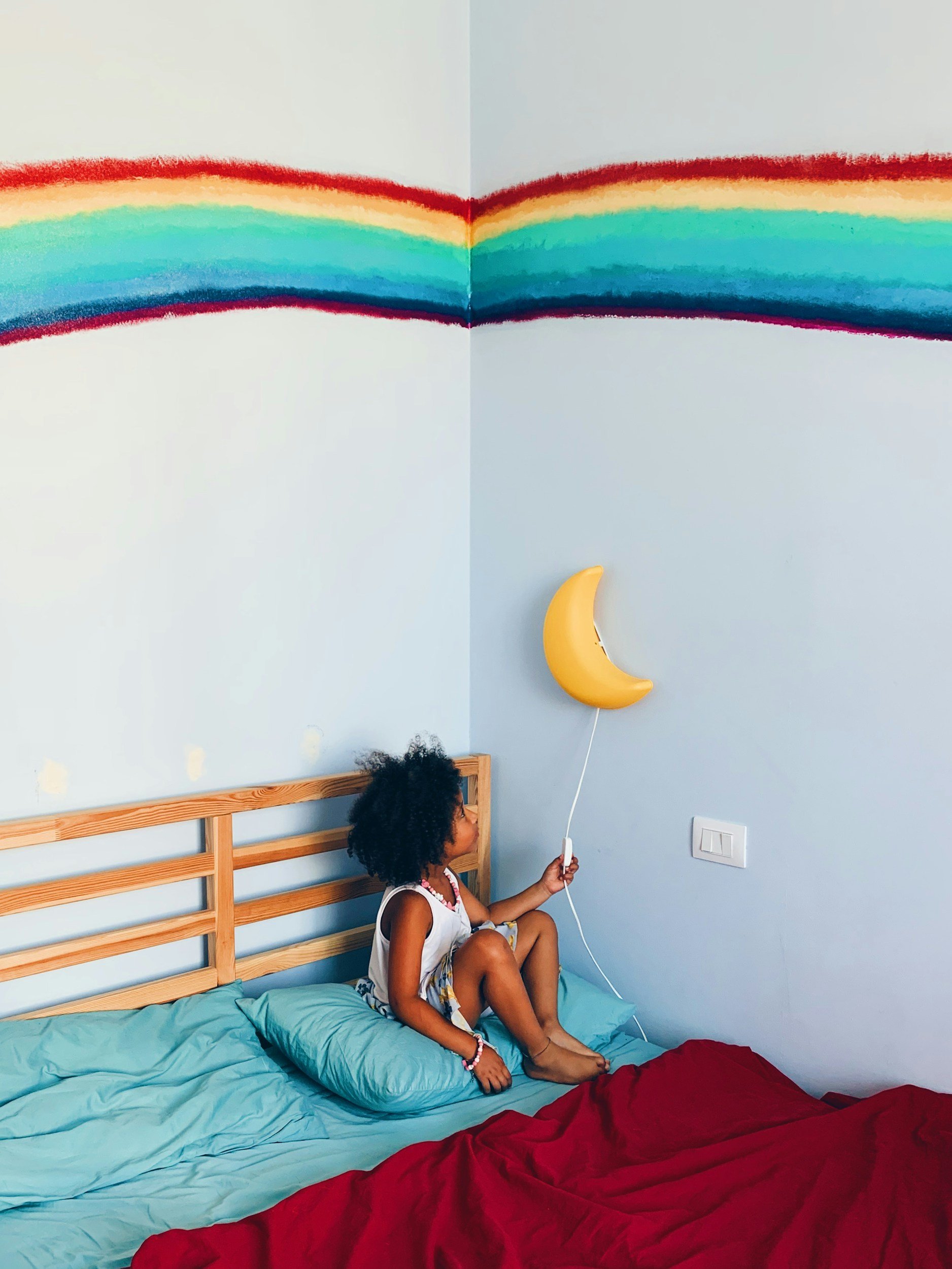 A young girl with curly hair, wearing a sleeveless top and patterned shorts, sitting on a bed with turquoise and red blankets, holding a yellow balloon in the shape of a crescent moon, near a blue wall with a rainbow mural and a yellow moon-shaped balloon.