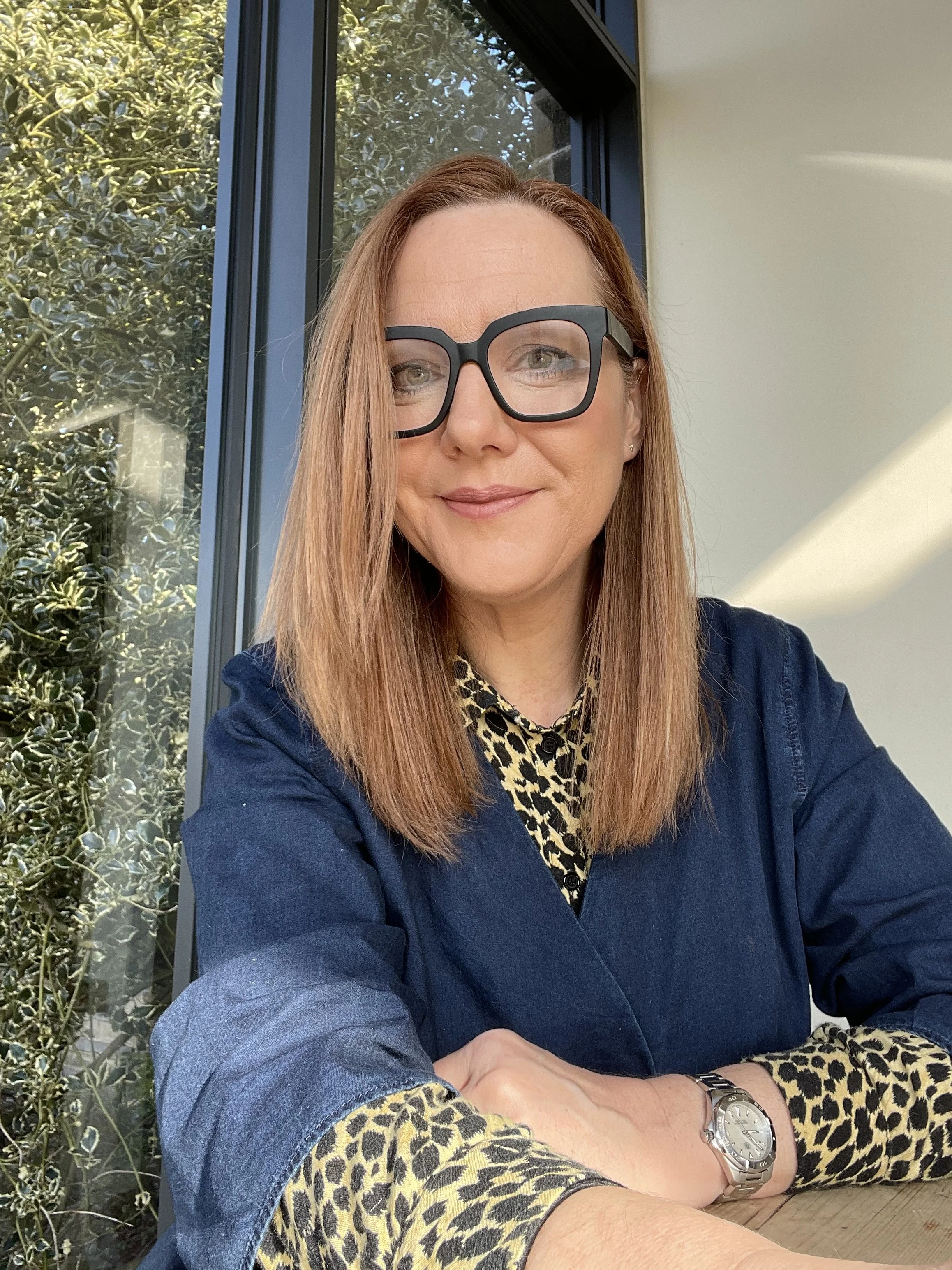 A woman with reddish-brown hair and glasses taking a selfie inside. She is wearing a leopard print top, a blue jacket, and a silver watch. She is sitting at a table with a window behind her, showing green foliage outside.