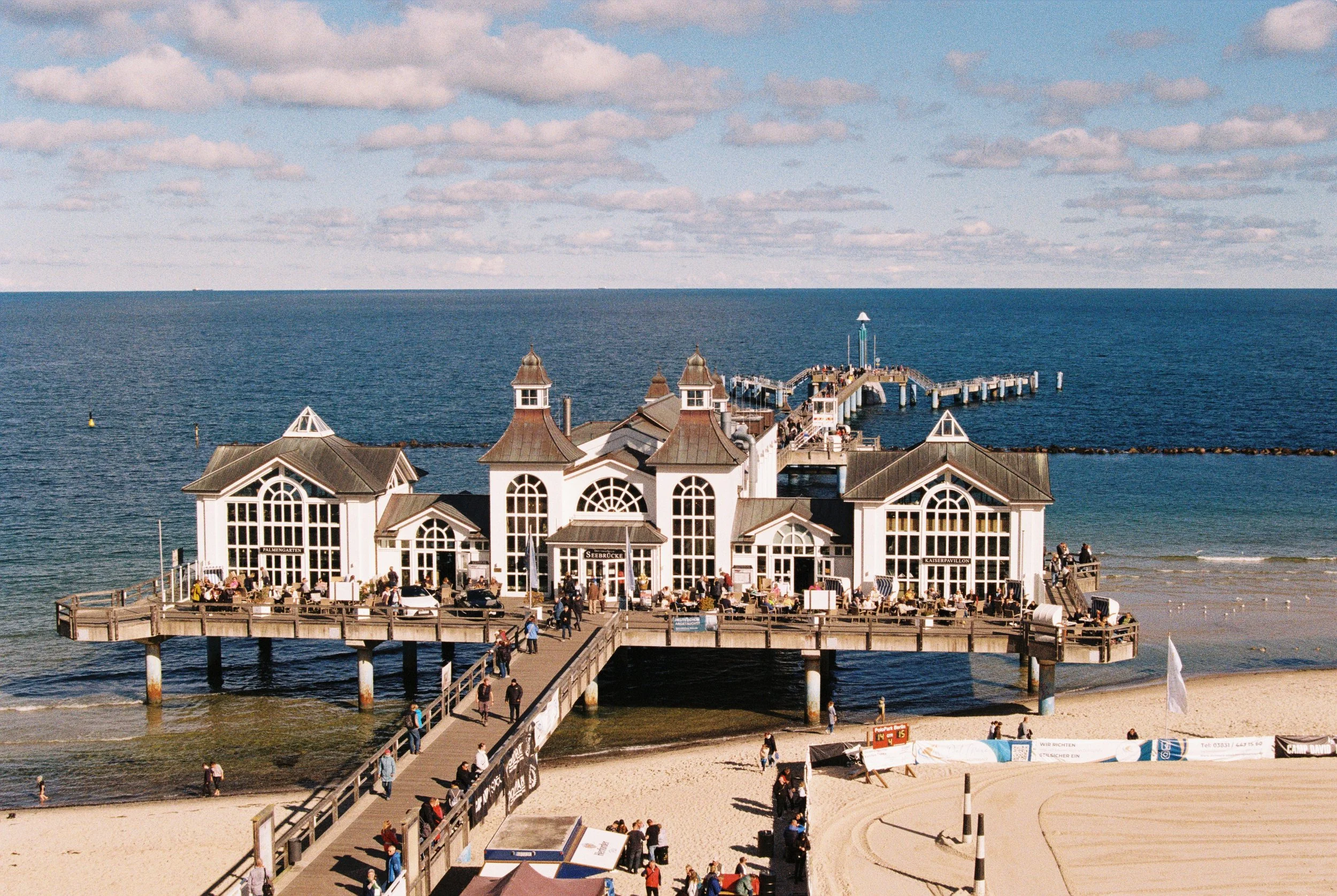 Das Bild zeigt ein Gebäude auf einem Steg, das zum Meer führt. Das Gebäude hat eine weiße Fassade mit großen Fenstern und mehreren Dachspitzen. Im Hintergrund ist der Ozean mit einem Pier, auf dem Menschen spazieren gehen, sowie eine bewölkte Himmel. Es gibt auch Menschen, die am Strand und auf dem Steg unterwegs sind.