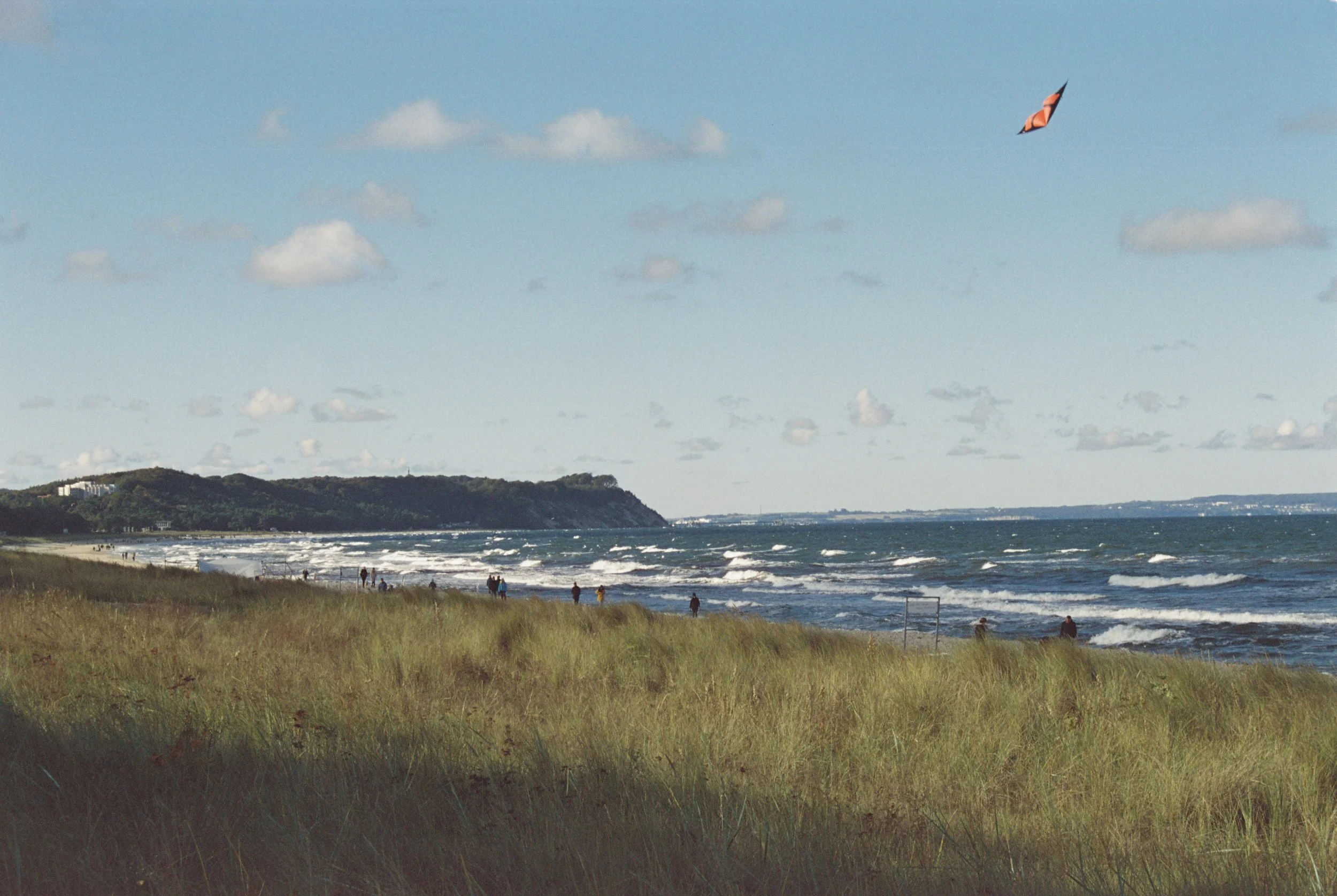 Strand mit Menschen, Wellen und einem Drachen am Himmel, Hügel im Hintergrund, bewölkter Himmel.