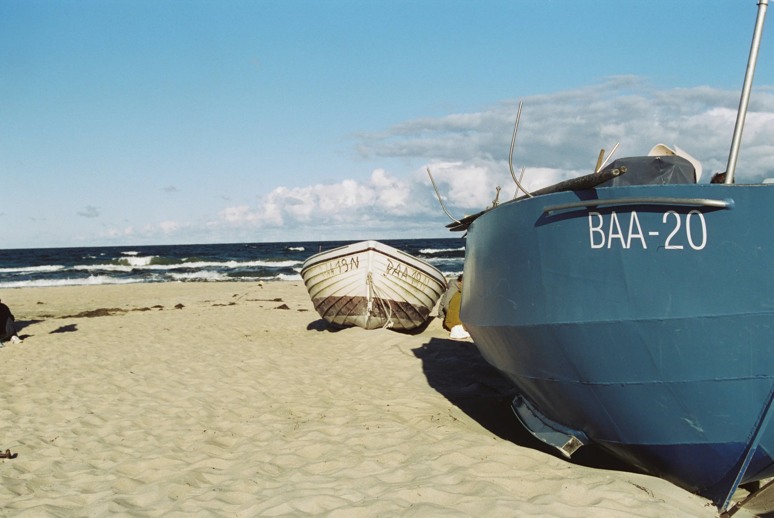 Zwei Boote am Strand, eines ist blau mit der Kennzeichnung 'BAA-20', das andere ist weiß mit schwarzen Linien. Der Strand ist sandig mit einigen Gegenständen verstreut, im Hintergrund das Meer und ein blauer Himmel mit wenigen Wolken.