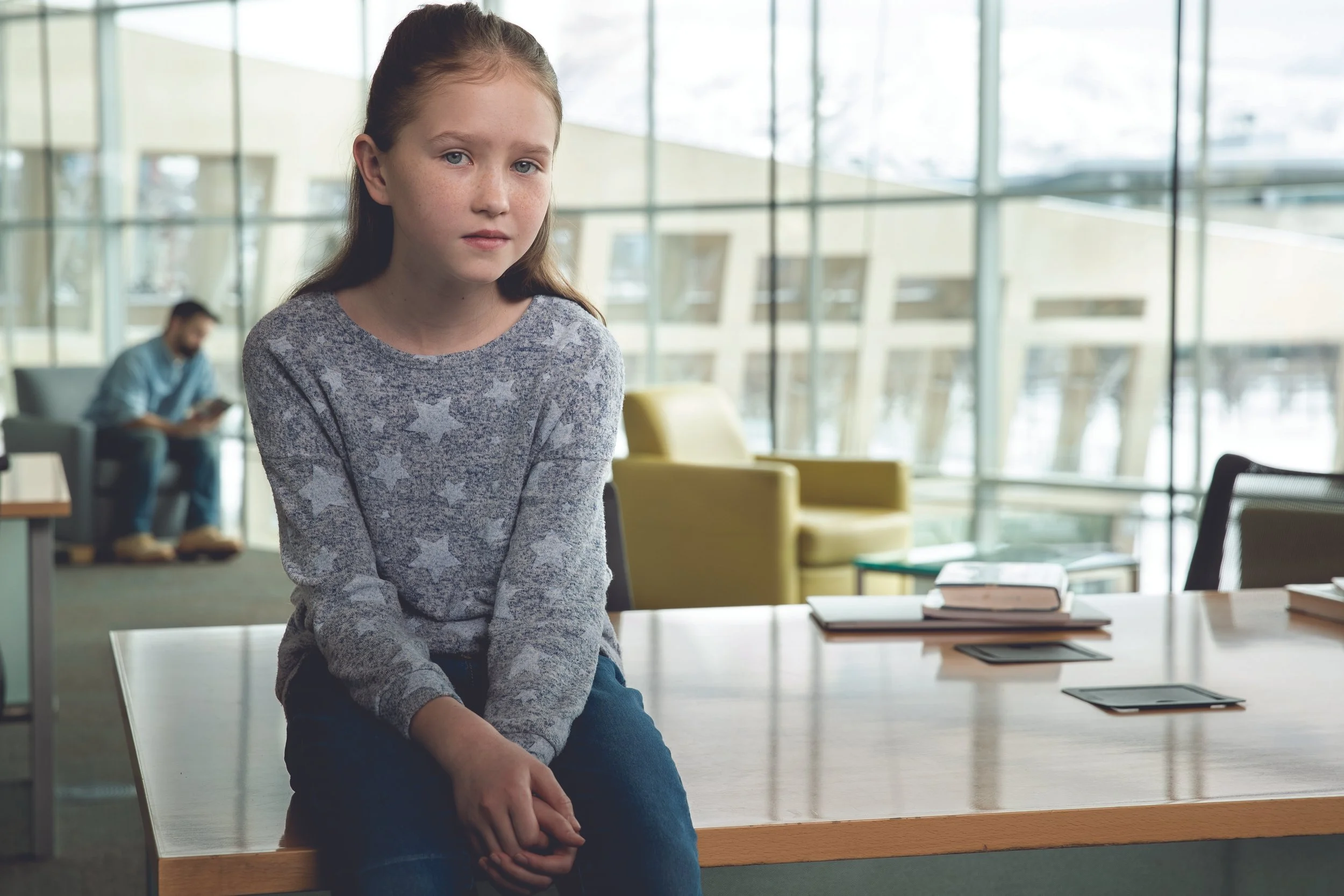 A young girl with light hair and freckles sitting on a desk in a modern indoor space, looking at the camera with a neutral expression. In the background, a man is sitting and reading a book near large glass windows.