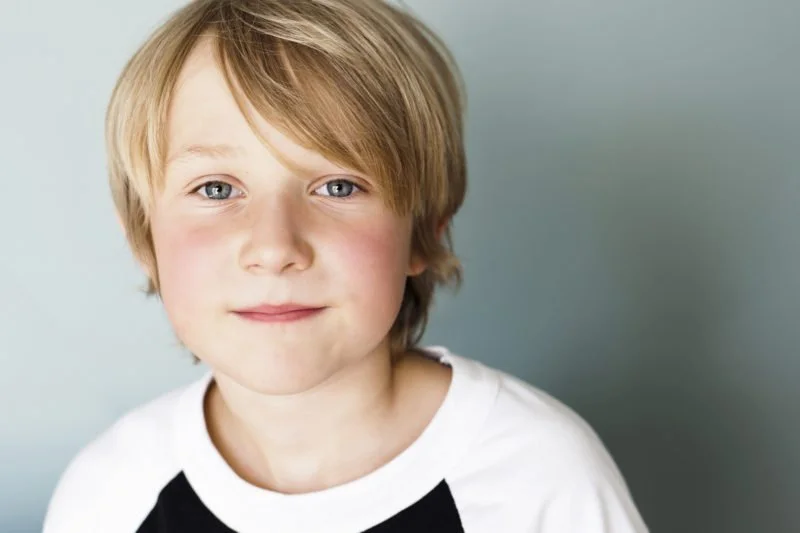 Close-up portrait of a young boy with blond hair and blue eyes wearing a white and black T-shirt, smiling softly against a plain light blue background.