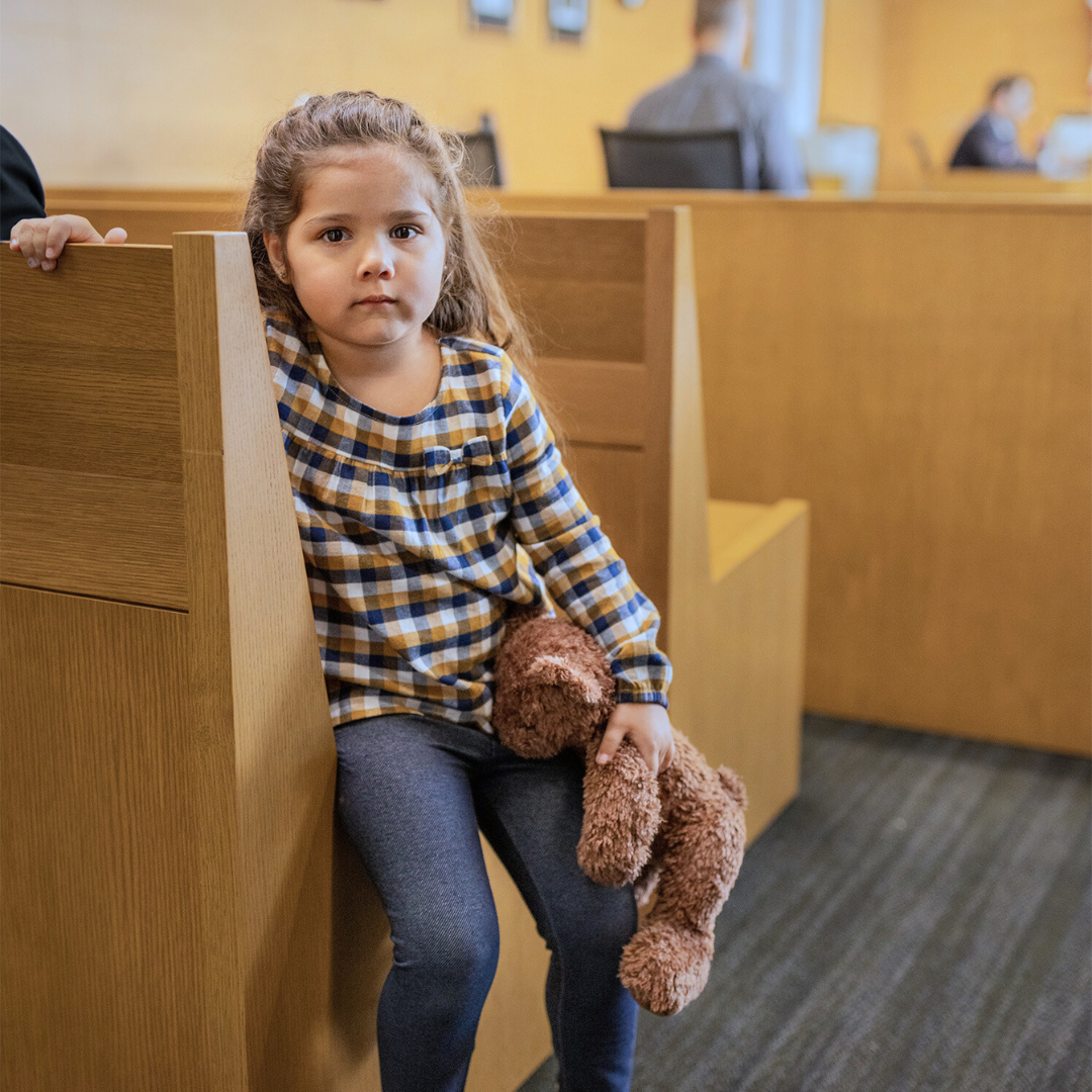 Young girl sitting on a wooden bench in a waiting area, holding a brown stuffed animal, with a serious expression.