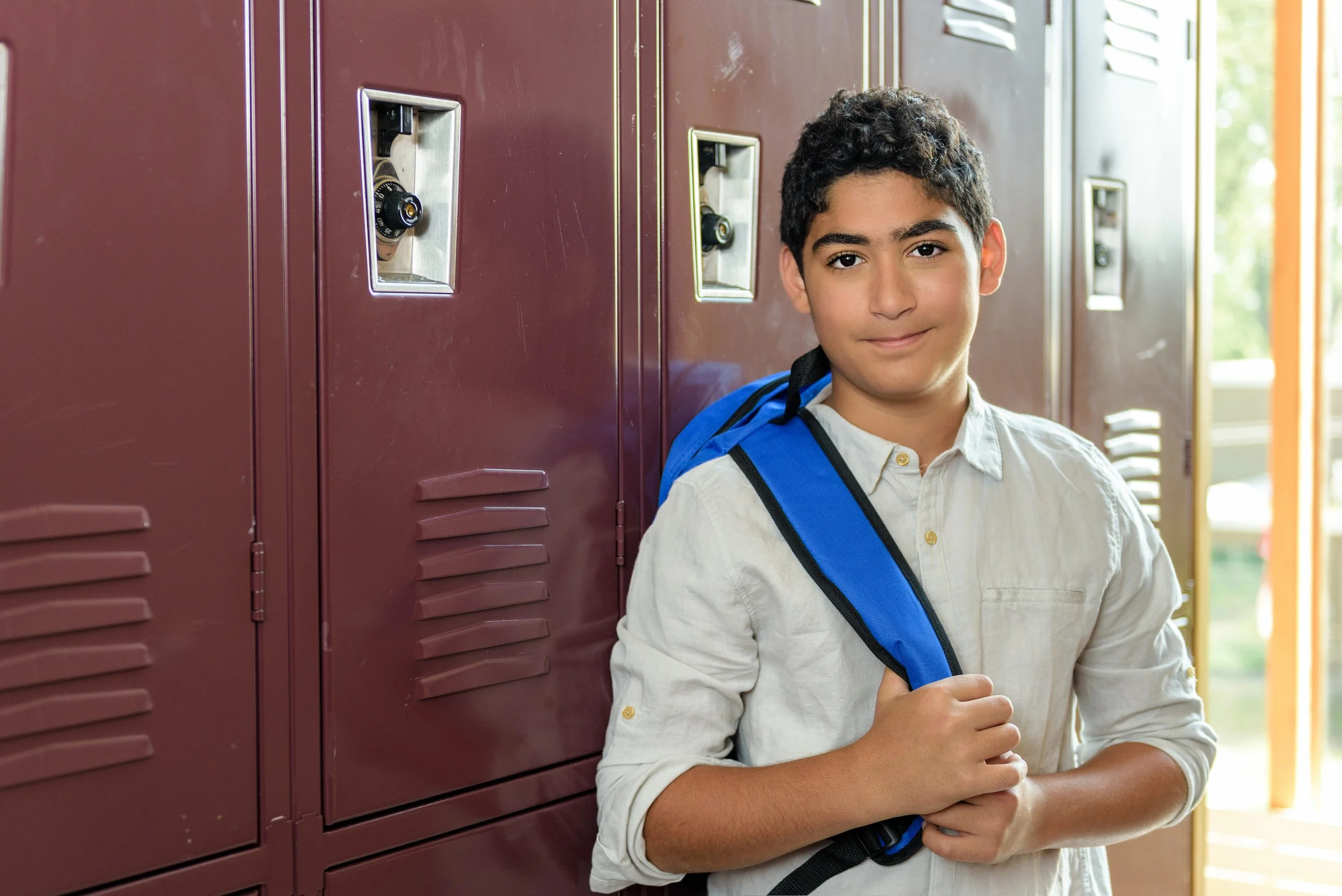 A young boy with curly black hair standing in front of maroon lockers in a school hallway, wearing a white shirt and carrying a blue backpack.