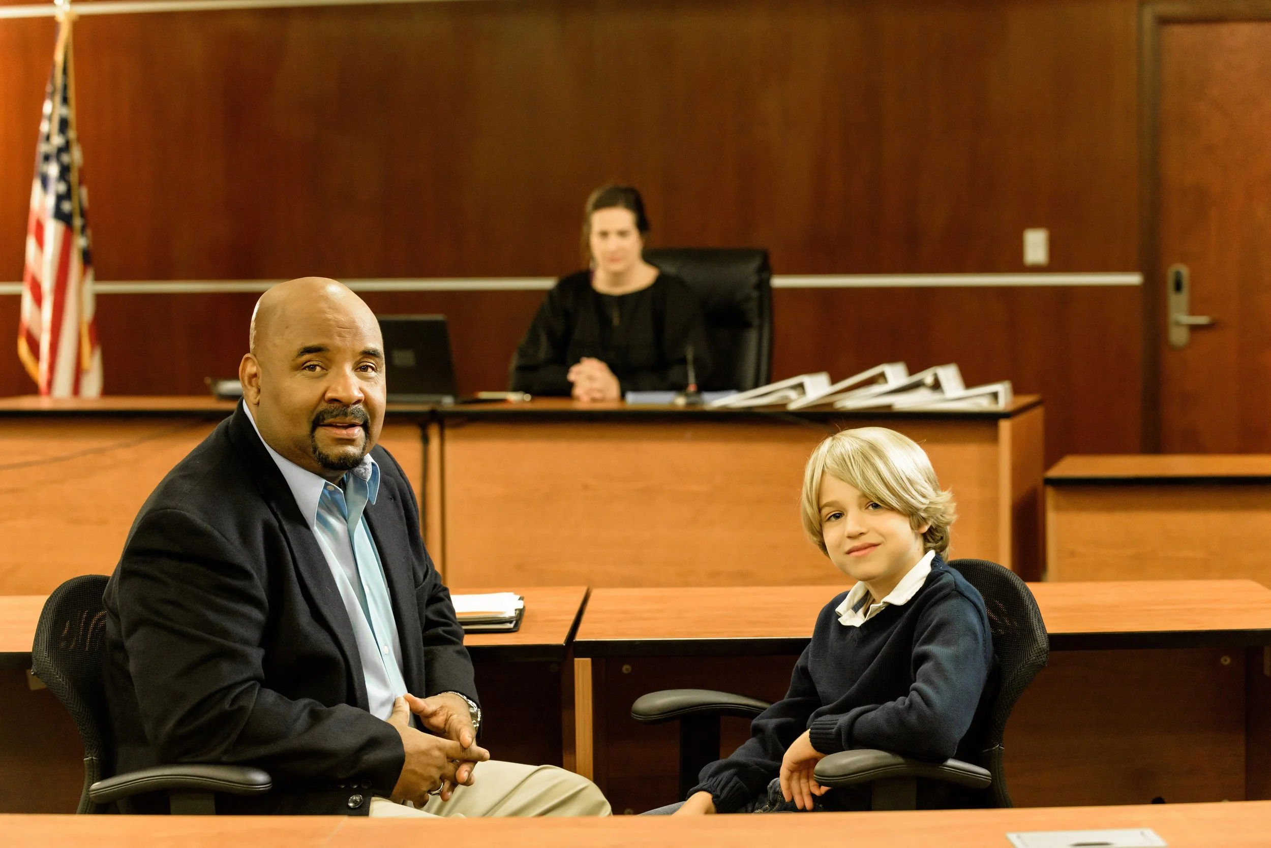 In a courtroom, a judge sits behind a bench with stacks of files, an American flag, and a woman in a black judicial robe. In the foreground, a man in a suit and a young boy shake hands, both seated at a table.