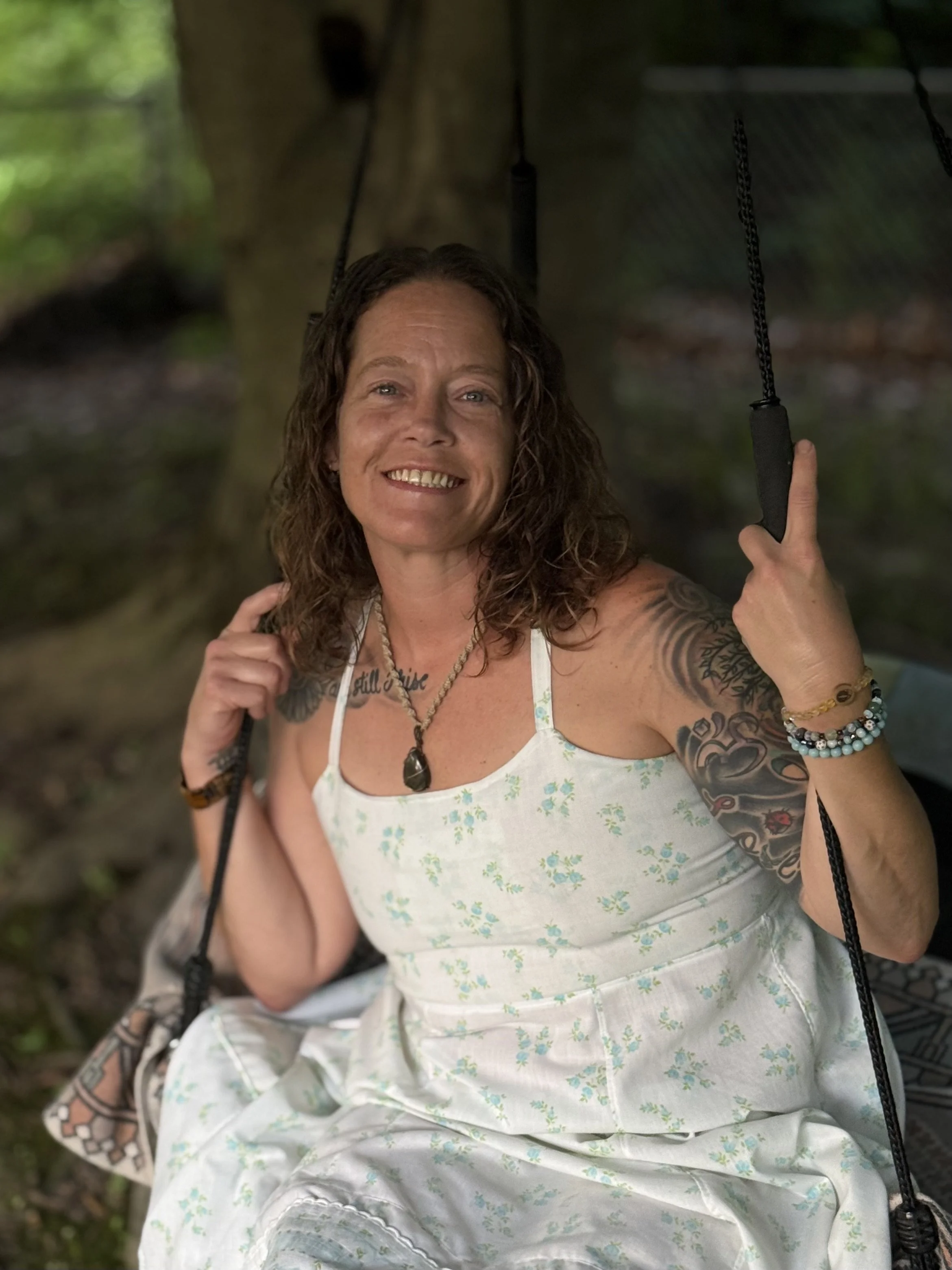 A woman with curly brown hair, tattoos on her arms, and wearing a white floral dress, smiling and sitting on a swing in a wooded area.