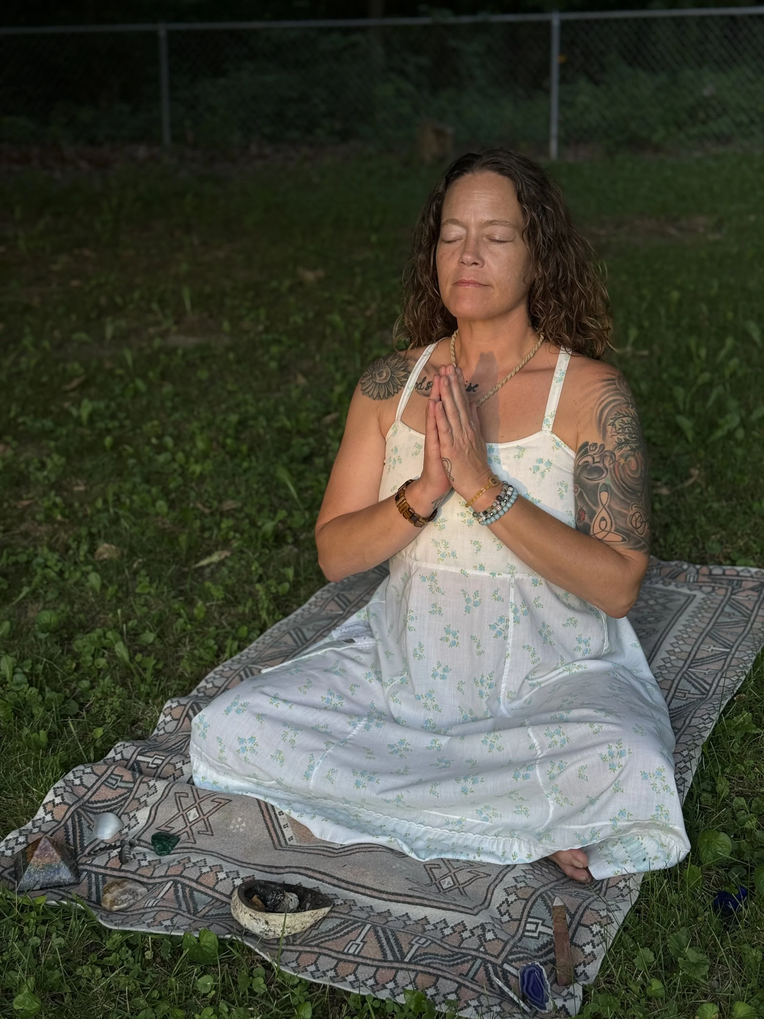 Woman with tattoos meditating outdoors on a blanket, with her hands in a prayer position and eyes closed.