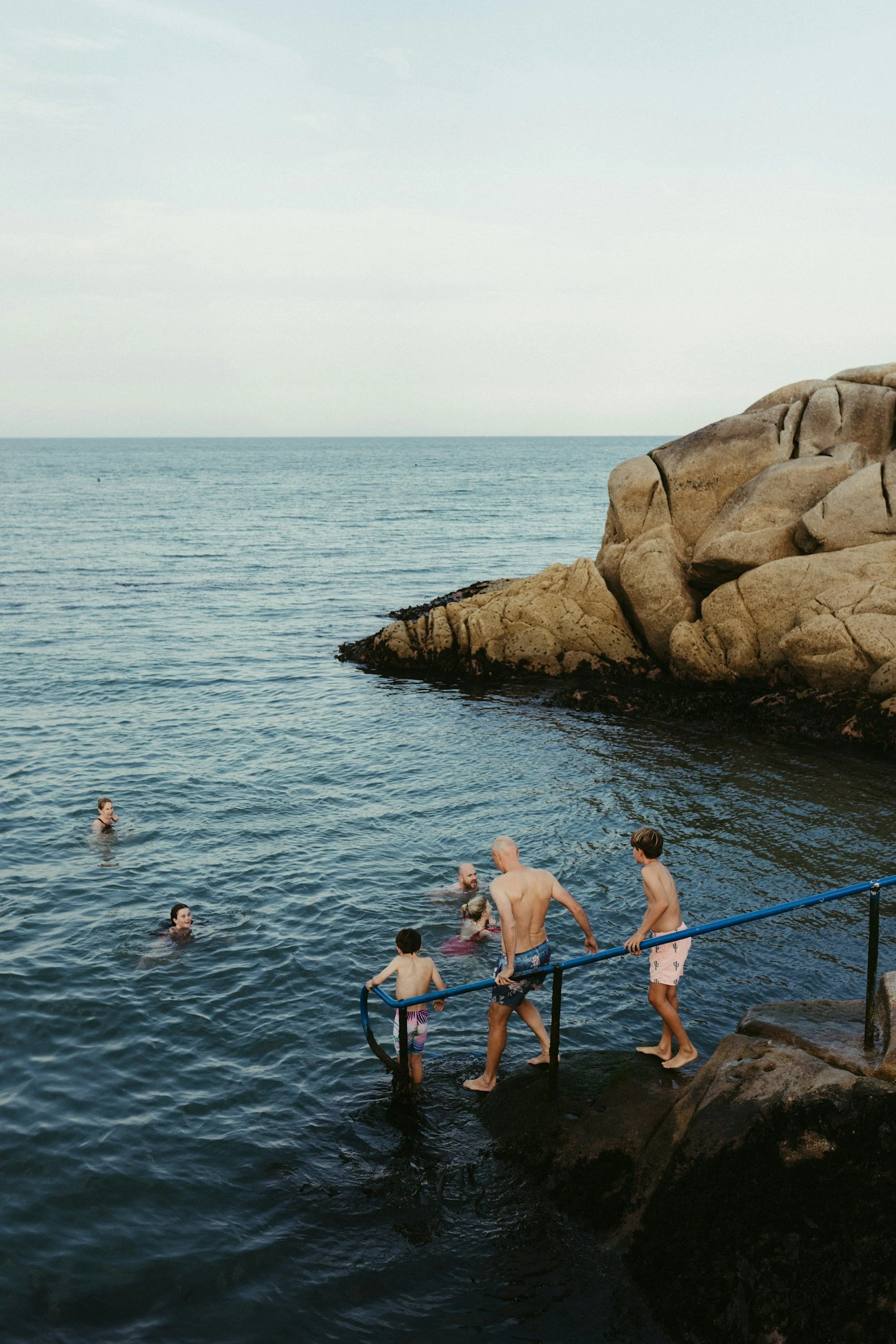 People swimming in the ocean near Dublin, and standing in the ocean near rocky shoreline with rocks and cliffs in the background.