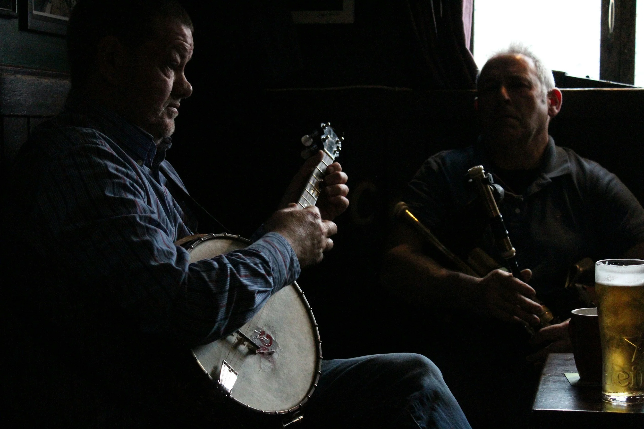 Two men sitting in an Irish bar, playing traditional musical instruments. One man is playing a banjo and the other is playing a clarinet. There are drinks on the table in front of them.