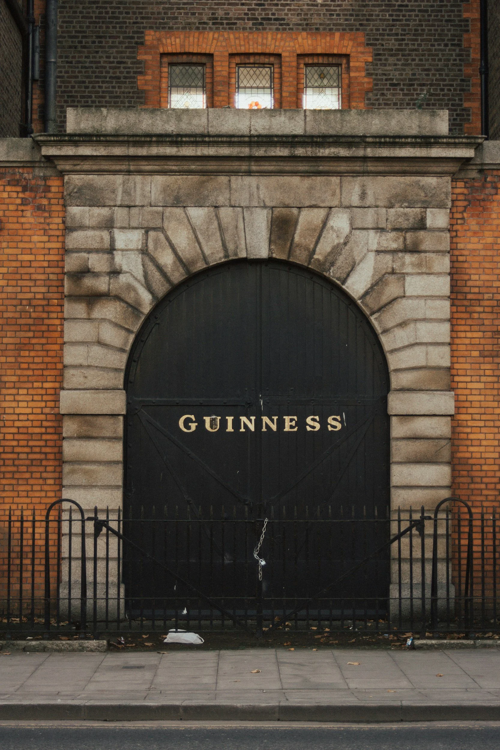 The facade of a brick building with three small window panes and a large arch doorway with a black door labeled 'GUINNESS'. The entrance is gated with a small black iron fence and chain.