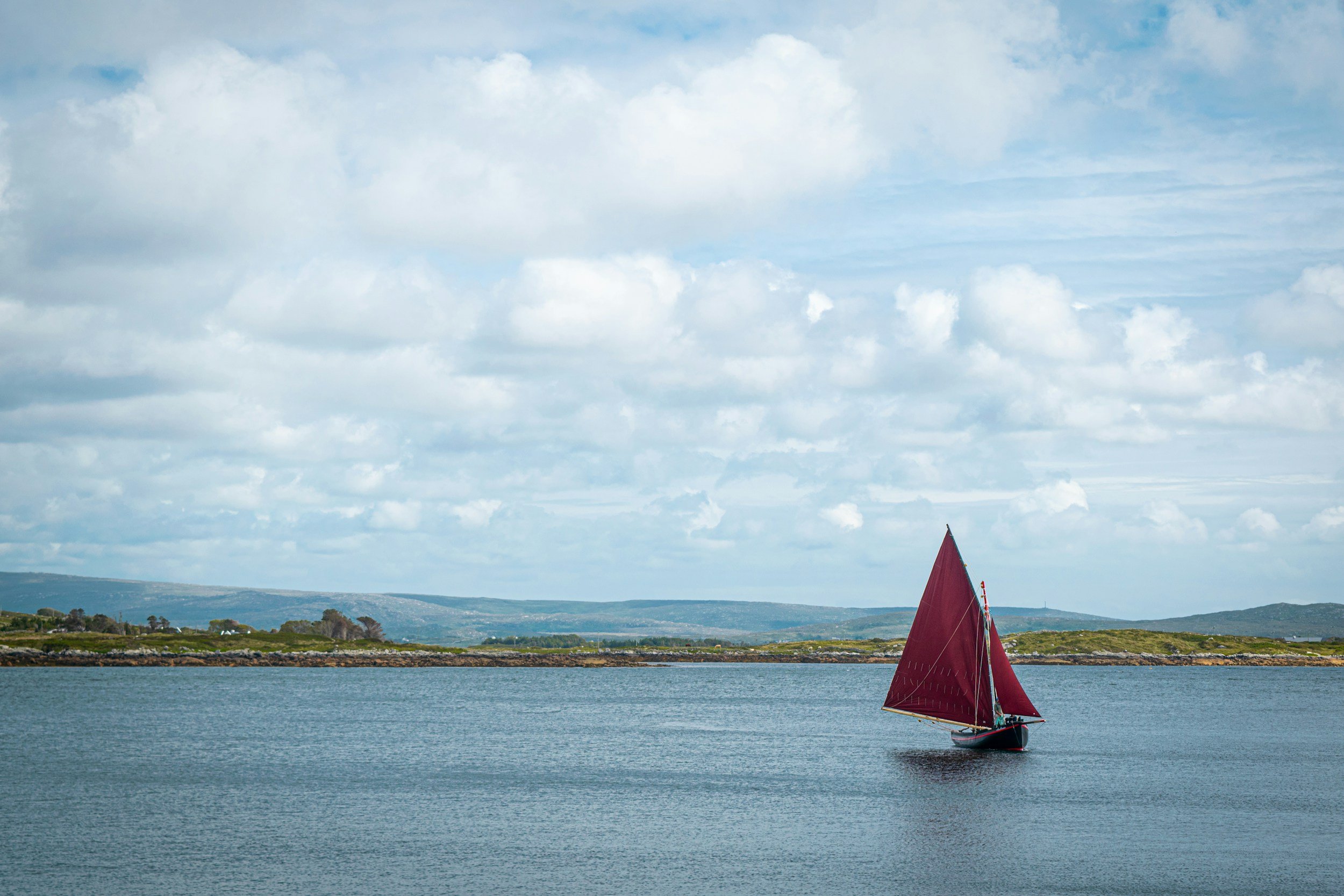 A red Galway Hooker sailboat with a triangular sail on a body of water, with a landscape of hills and a cloudy sky in the background.