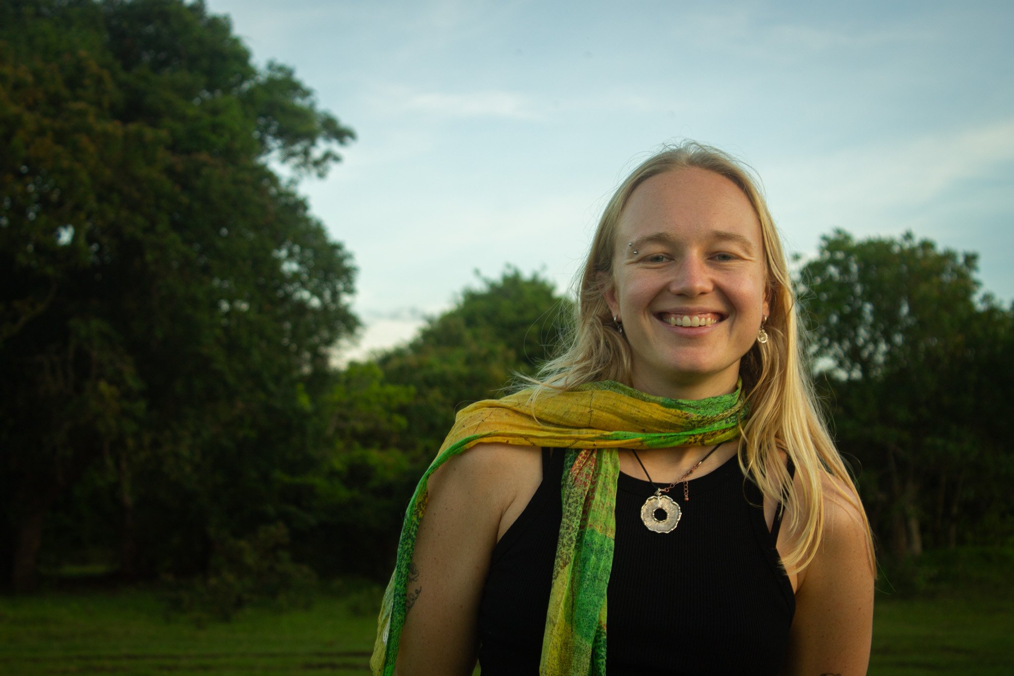 A woman with blonde hair wearing a black top and a green patterned scarf smiling outdoors with trees and a cloudy sky in the background.