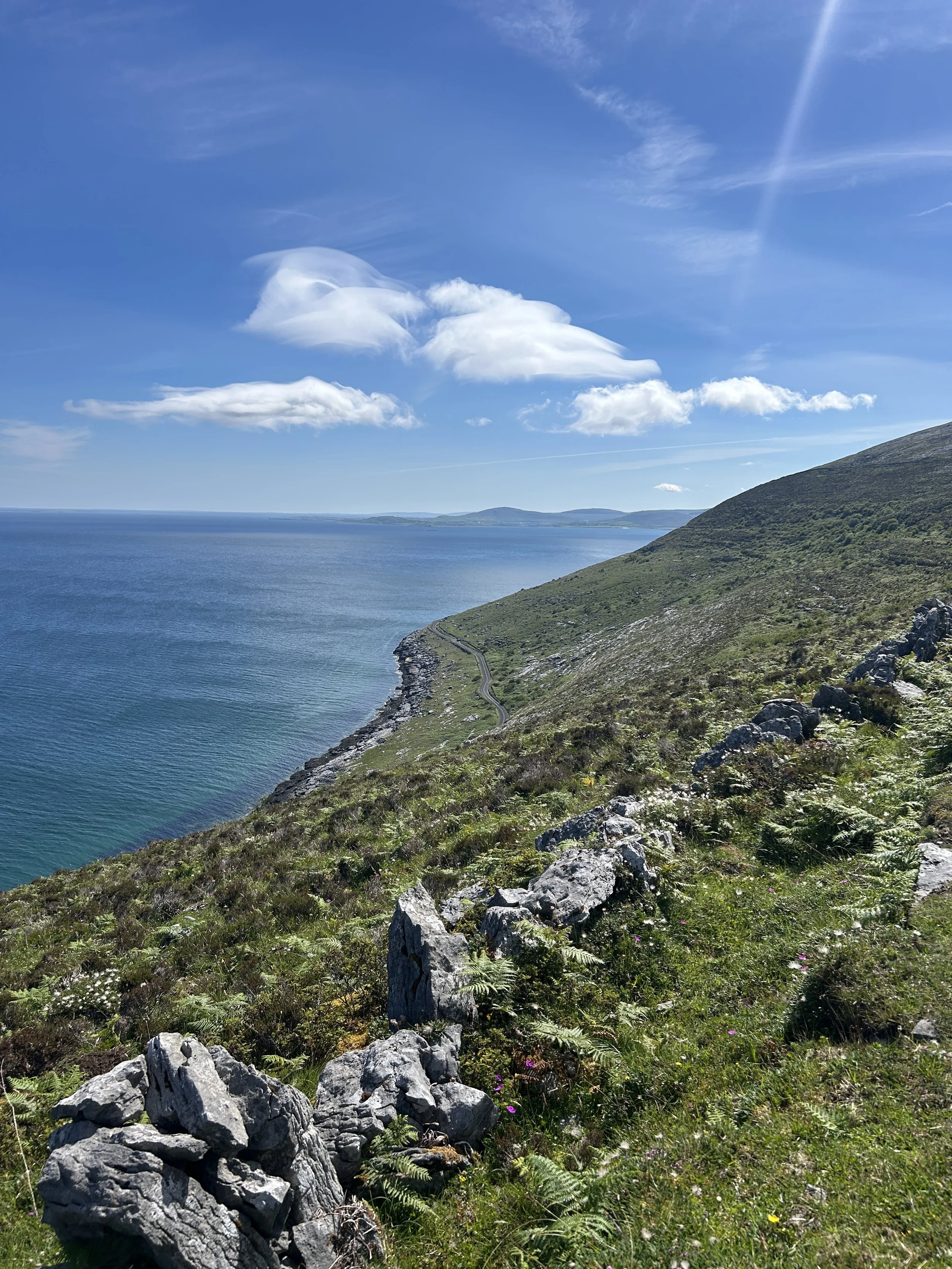 Scenic view of The Burren and the ocean with rolling green hills and rocky terrain along the coast under a blue sky with white clouds.