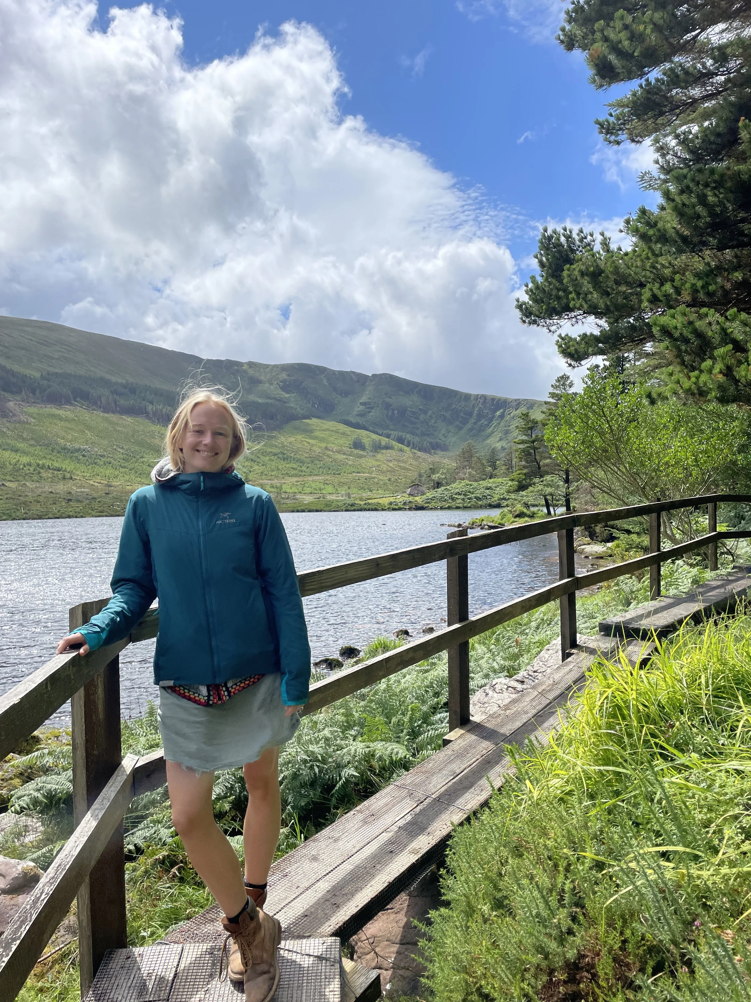 A woman standing on a small bridge near a lake, surrounded by green plants and trees, with mountains and a blue sky with clouds in the background.