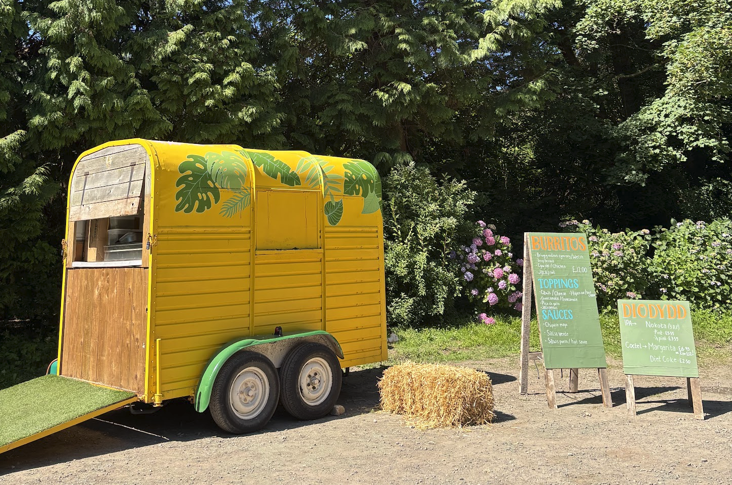 A yellow food trailer with green leaf decorations, parked next to a hay bale, with colorful flower bushes and greenery in the background. Two green chalkboard signs list burrito and drink options.