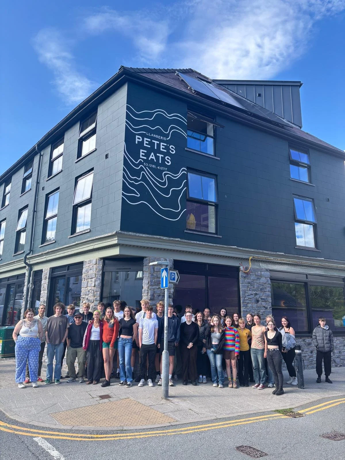 A group of young people standing outside a modern building with a sign that reads 'Llanberis Pete's Eats' and a mountain top map design, under a blue sky with some clouds.