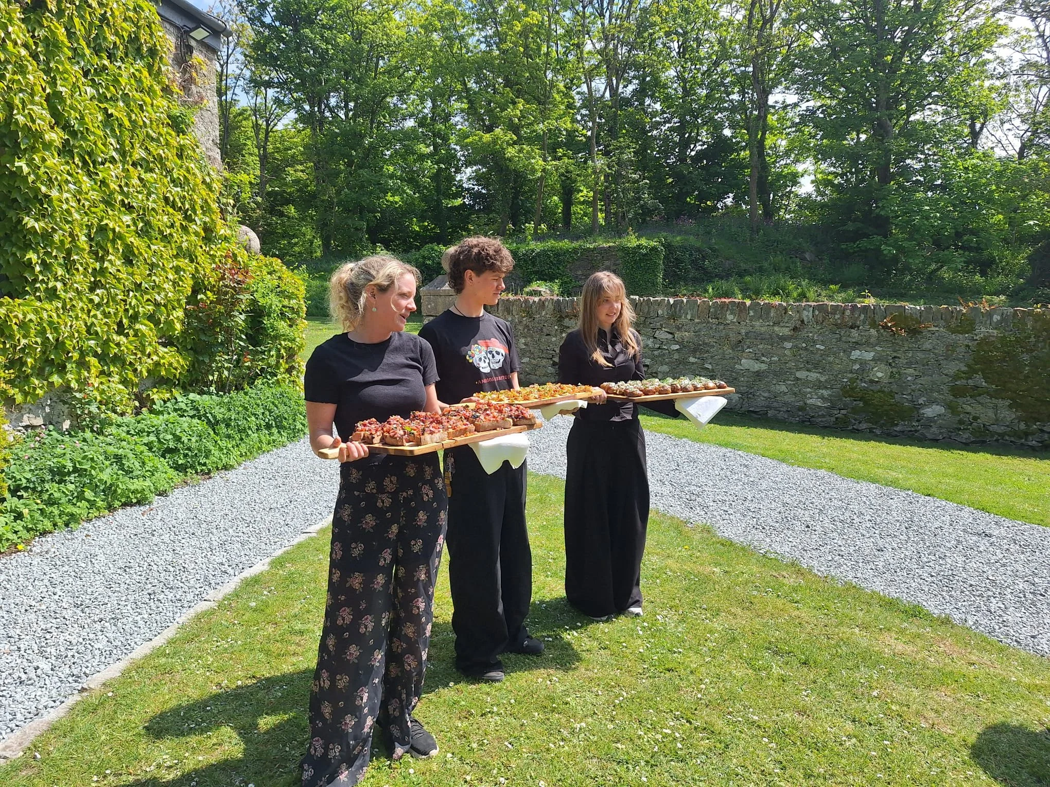 Three women standing outdoors on a gravel path beside a stone wall, with lush green trees and grass in the background. Two women are carrying trays of food, possibly pastries or desserts, and are dressed in black outfits.
