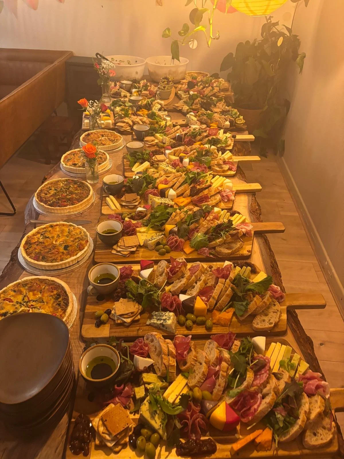 A long wooden table filled with assorted cheeses, sliced meats, crackers, bread, salads, pizzas, and small bowls of olive oil, surrounded by potted plants and a warm, cozy atmosphere.