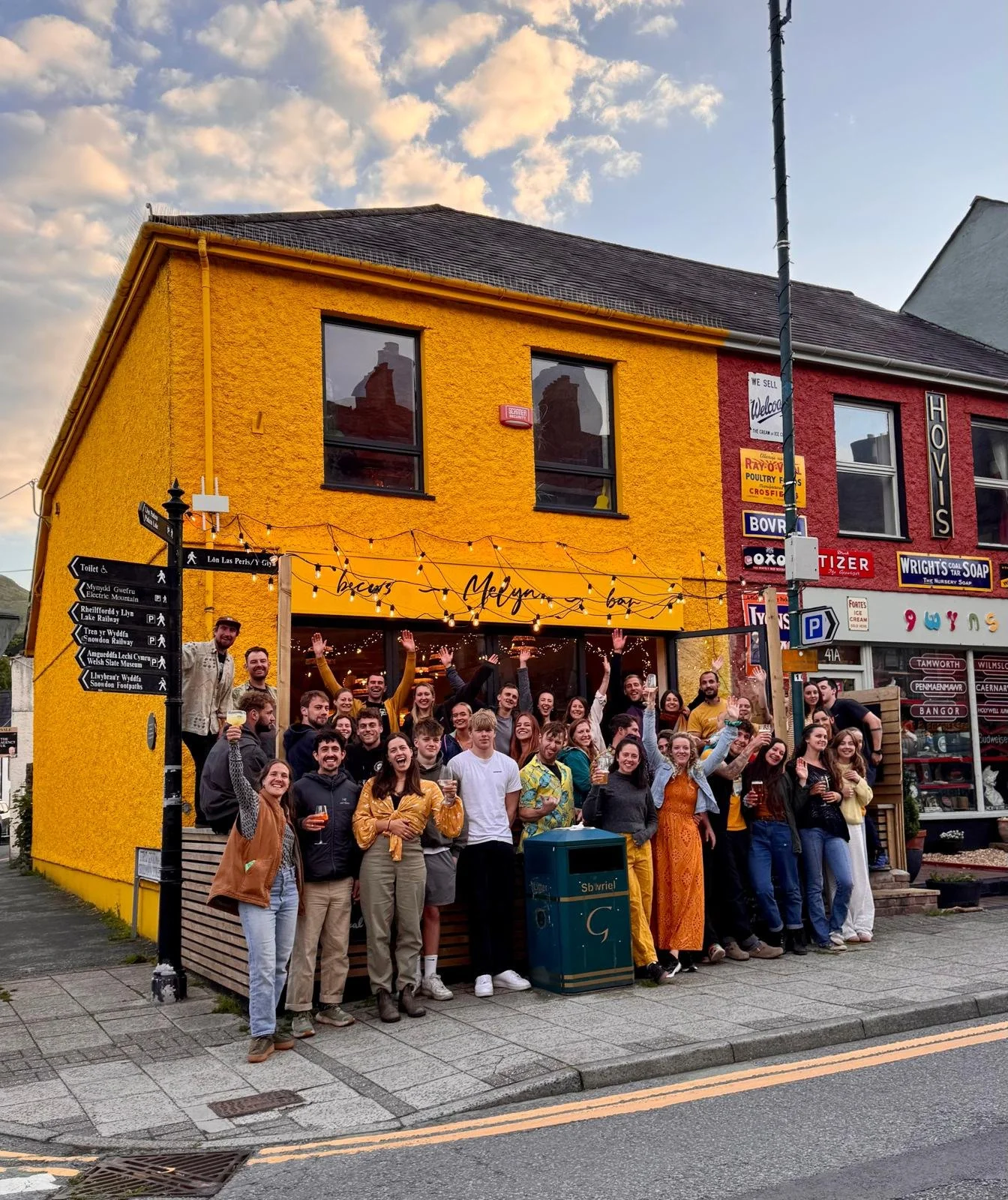A group of people gathered outside a yellow building with string lights, some holding drinks and waving. The building has signs and a mural, with a cloudy sky above.