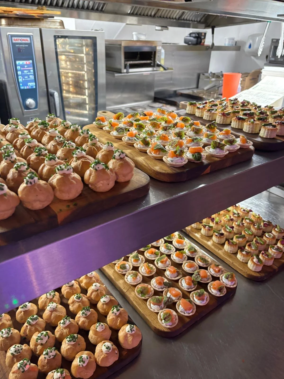 Assorted appetizers on wooden boards in a restaurant kitchen, including small round pastries topped with cream and herbs, and toasted bread bites with vegetables and herbs, arranged on a stainless steel counter.