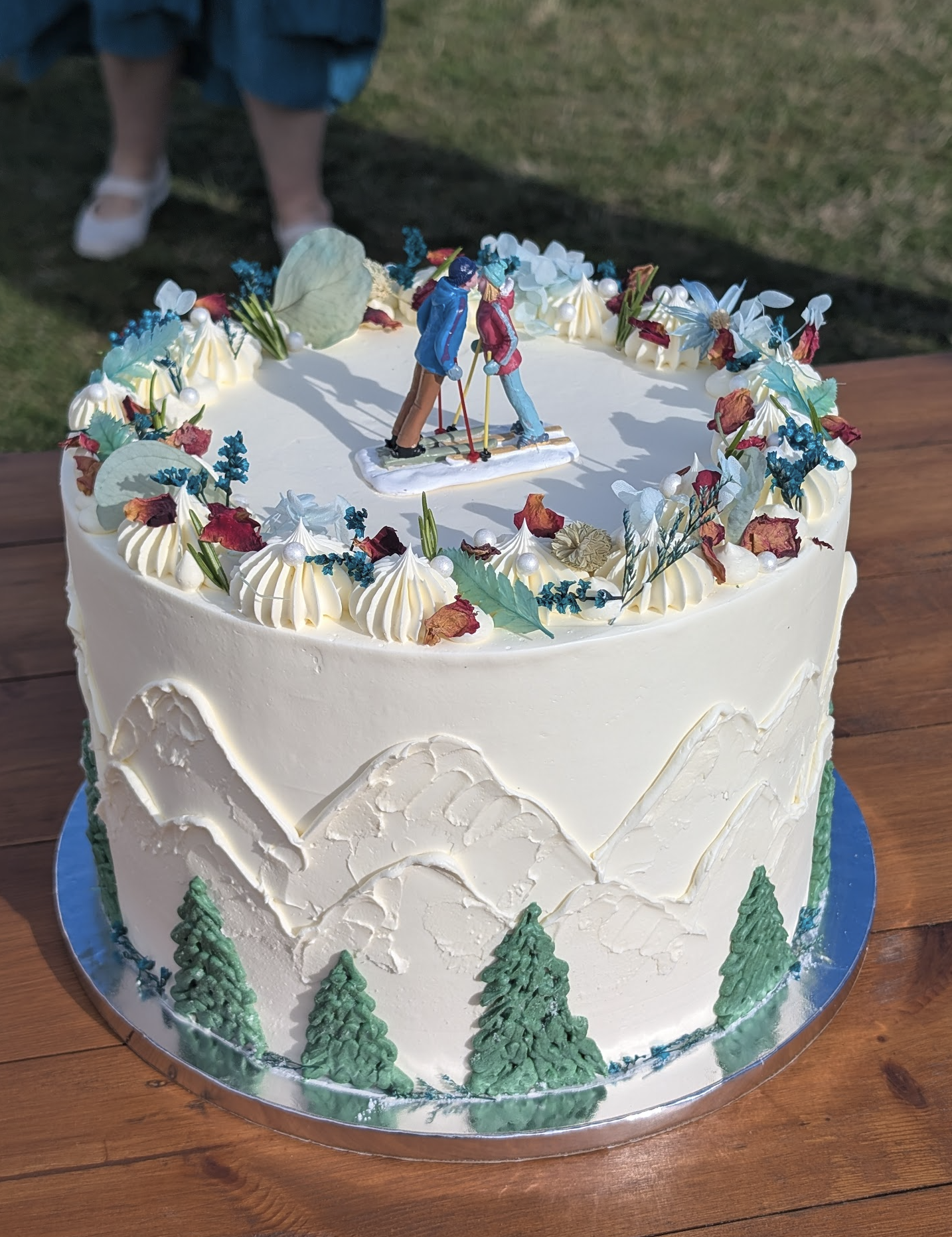 A white cake decorated with small green trees at the bottom, white icing swirls with edible pearls and colorful dried flowers on top, and a miniature skier figurine as a topper.