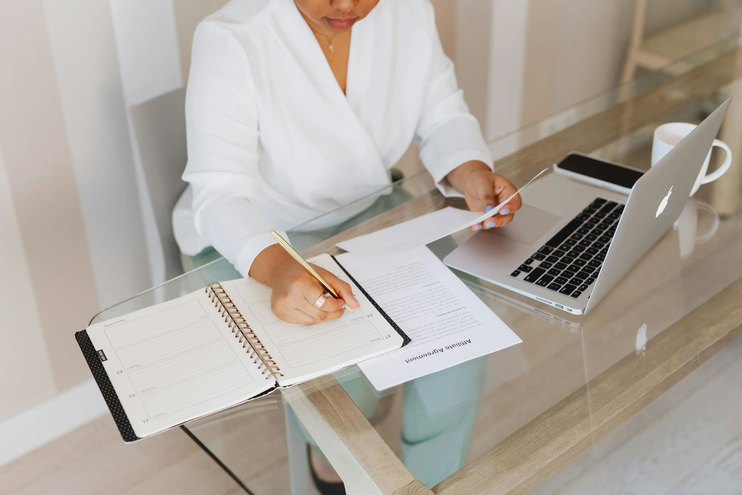 Person sitting at a desk with a laptop, writing in a planner and holding a piece of paper.