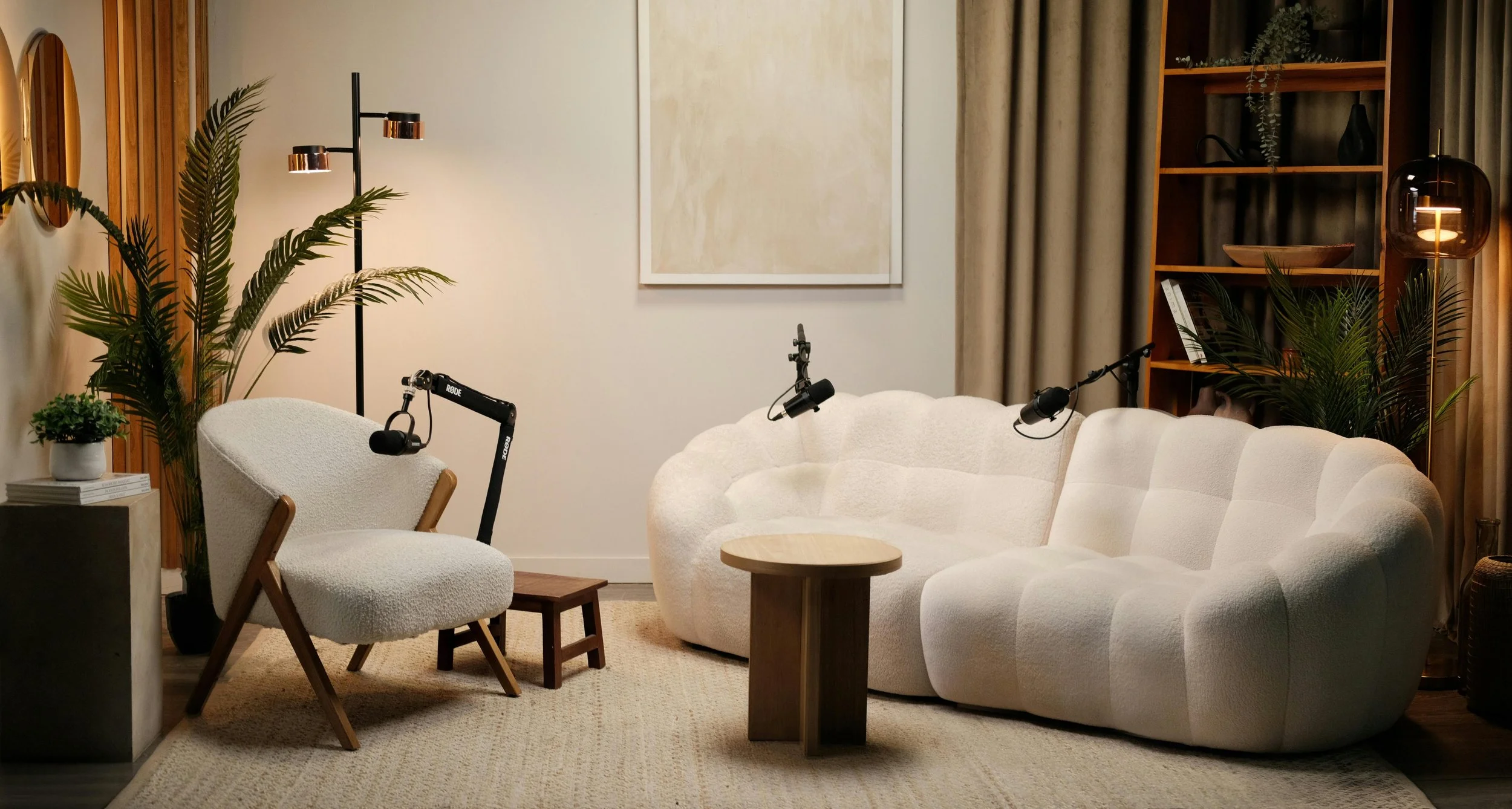 A cozy living room with white textured sofas, a cream armchair, small wooden tables, potted plants, a bookshelf, a floor lamp, and wall art.
