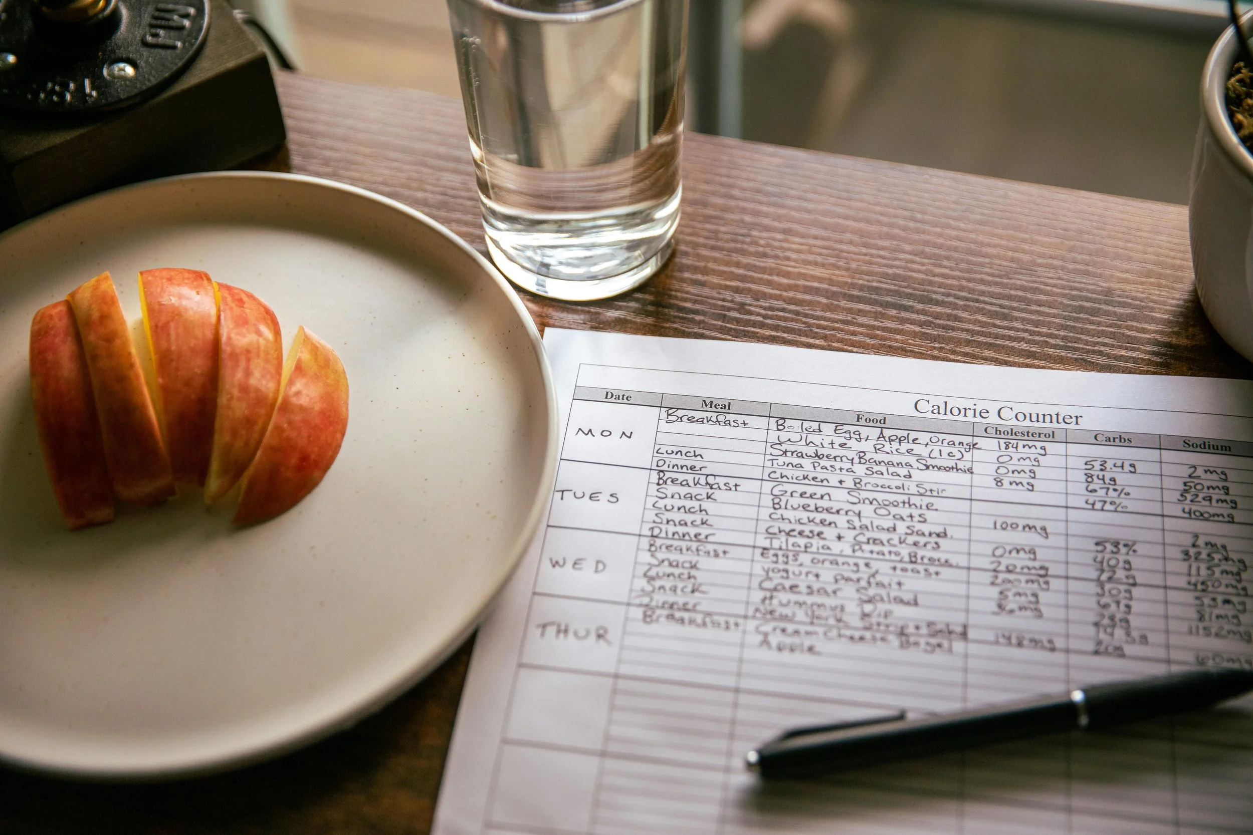A breakfast plate with sliced apple, a glass of water, a sheet of paper with a calorie counter, a pen, a clock, and a bowl of cereal on a wooden surface.