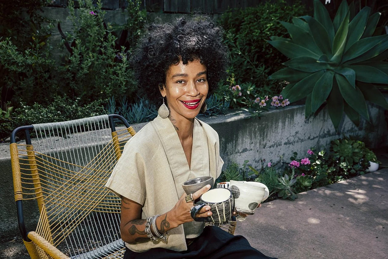 Woman with curly hair smiling, sitting outdoors in a garden, holding ceramic cups.