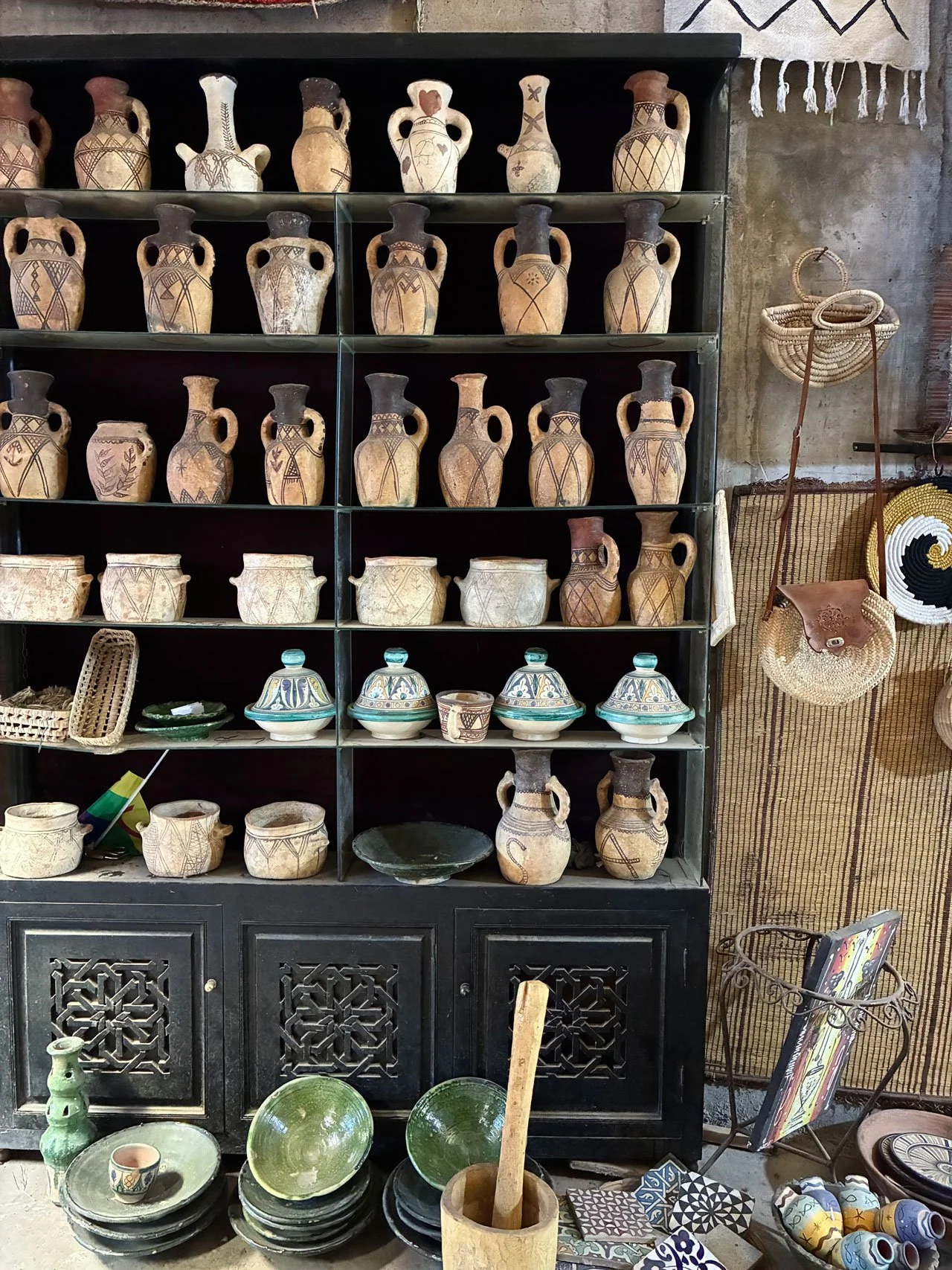 Display of traditional pottery and ceramic items on a black shelf. The items include vases, bowls, and patterned pottery, along with some woven baskets and decorative accessories on the side and bottom.