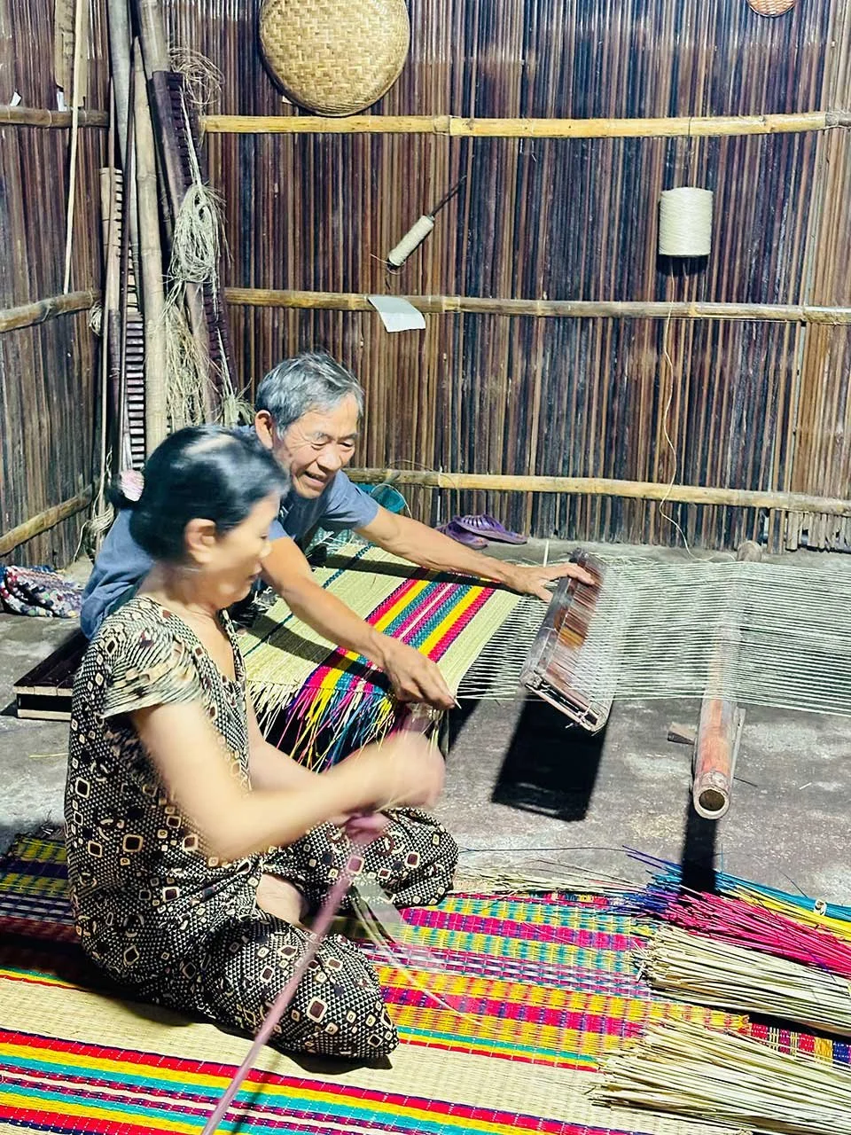 Two elderly people weaving colorful textiles on traditional wooden looms inside a bamboo-walled room.
