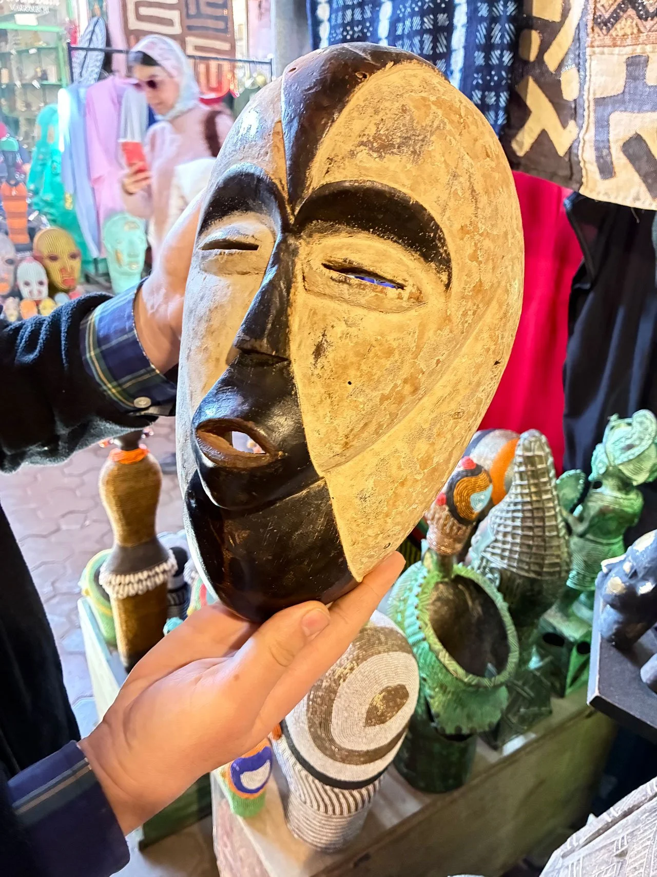 A wooden African mask with a split face design, one half painted black and the other natural wood color, being held by a person in a marketplace with colorful traditional crafts and masks in the background.