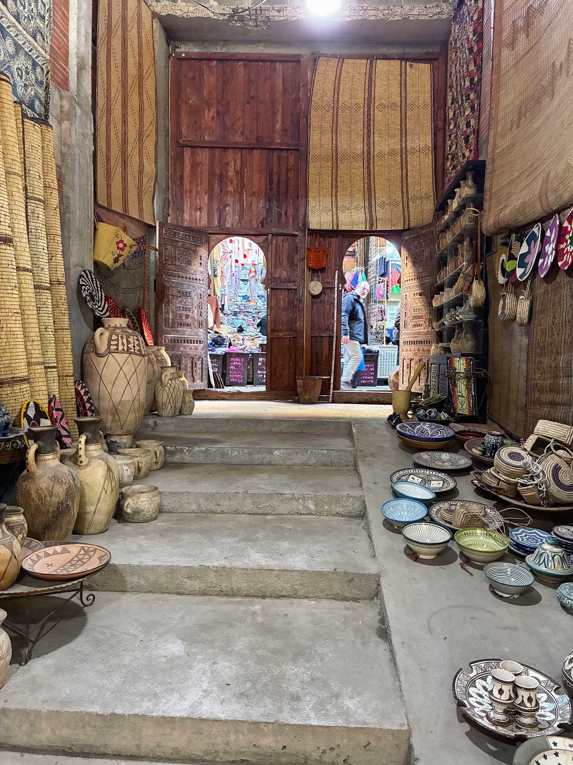 A collection of pottery and woven baskets displayed in a rustic shop with wooden doors and a bustling outdoor market visible through the open doorways.