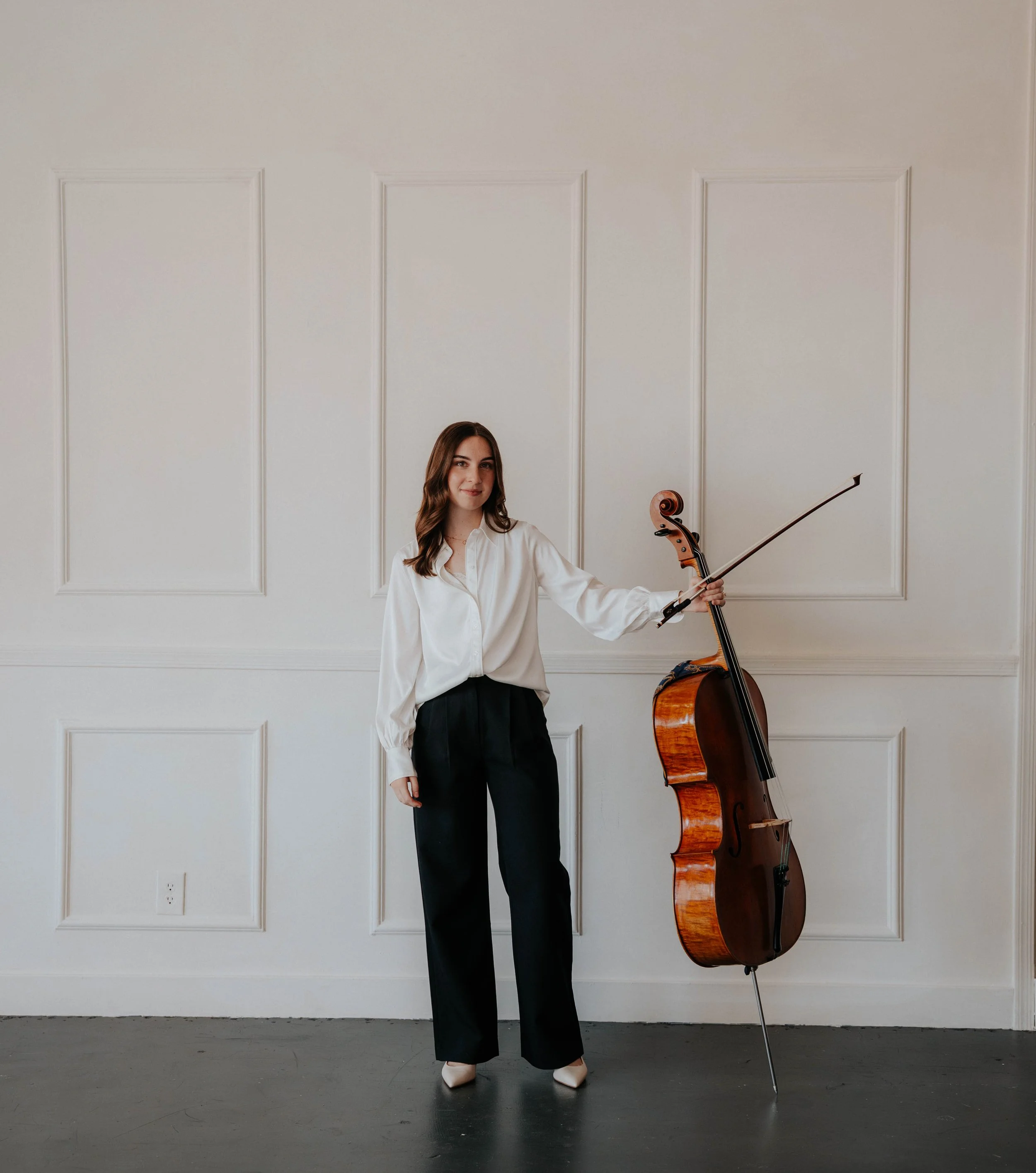 A young woman with brown hair wearing a white shirt and black pants, standing in front of a white paneled wall, holding a cello.