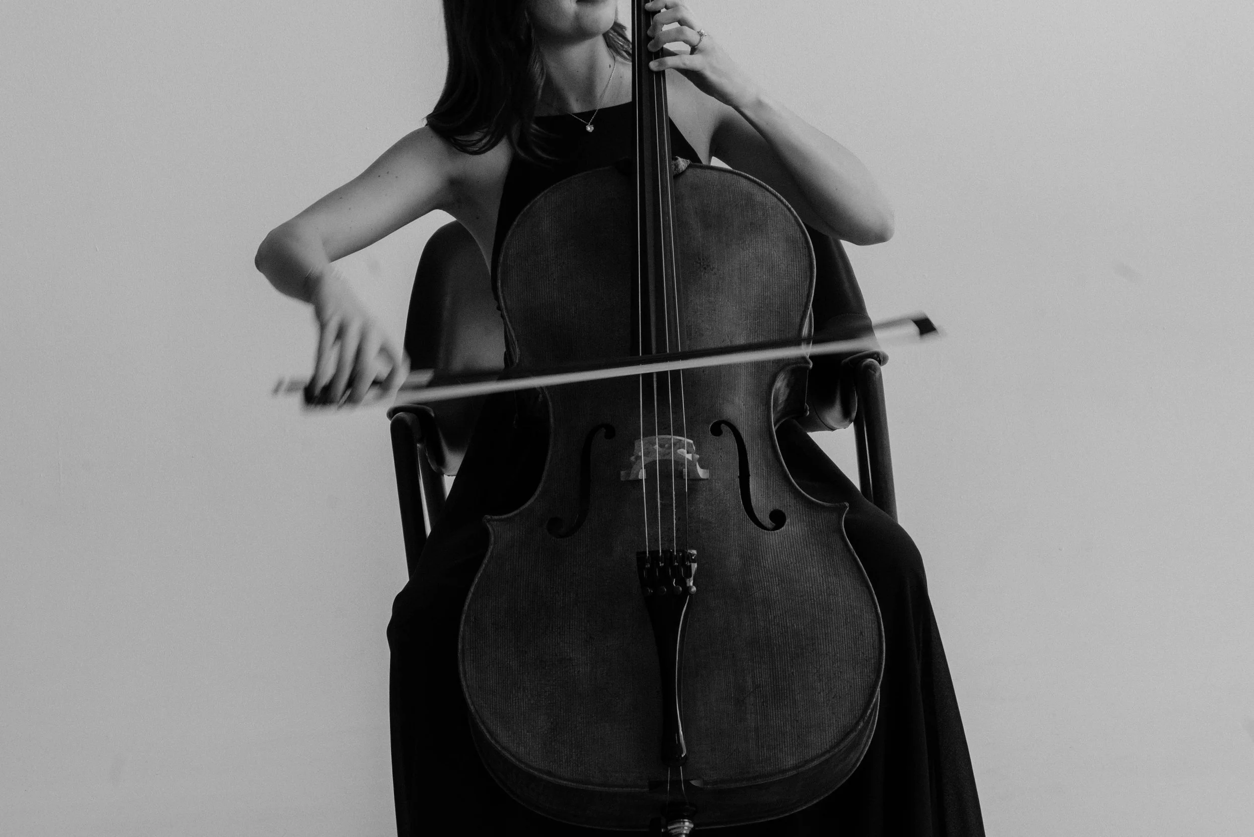 A woman playing a cello, sitting on a chair, with a plain light-colored wall in the background.