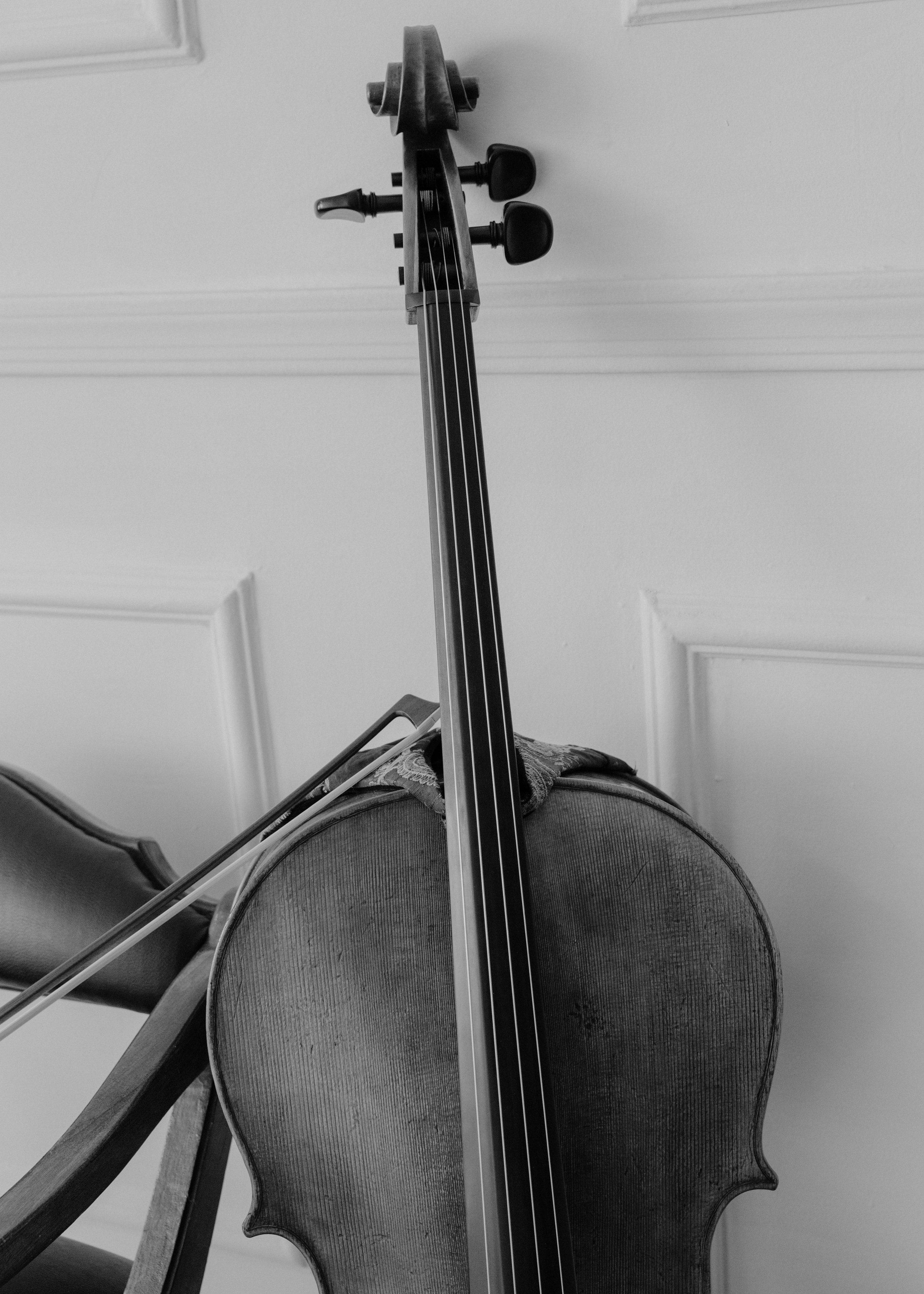 A black and white photo of a cello resting on a chair, with the camera angle from below, capturing the scroll, pegs, and strings of the instrument.