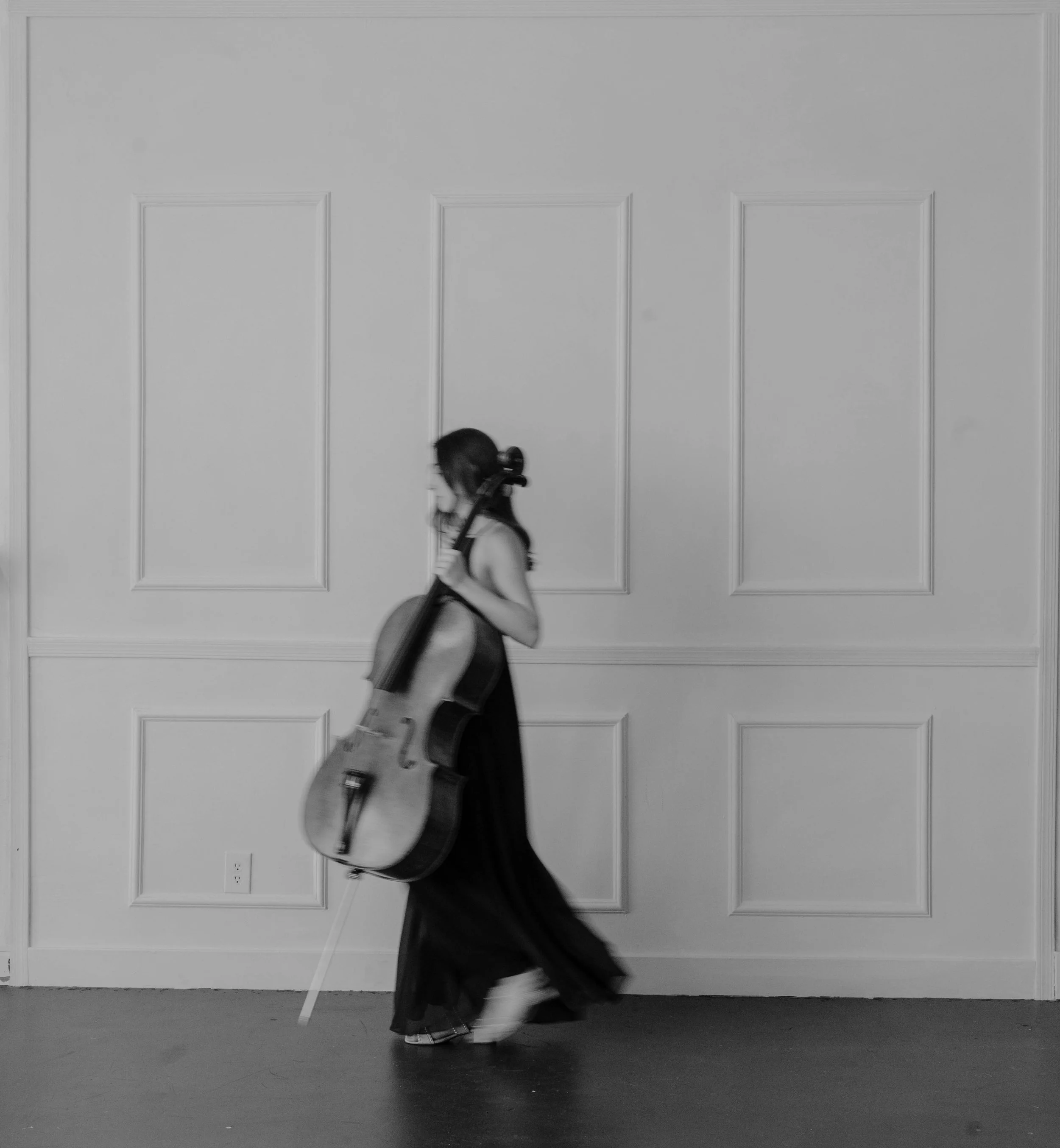 A woman with long hair, wearing a dress, is walking across an indoor space while carrying a cello. The background features a plain wall with paneling.